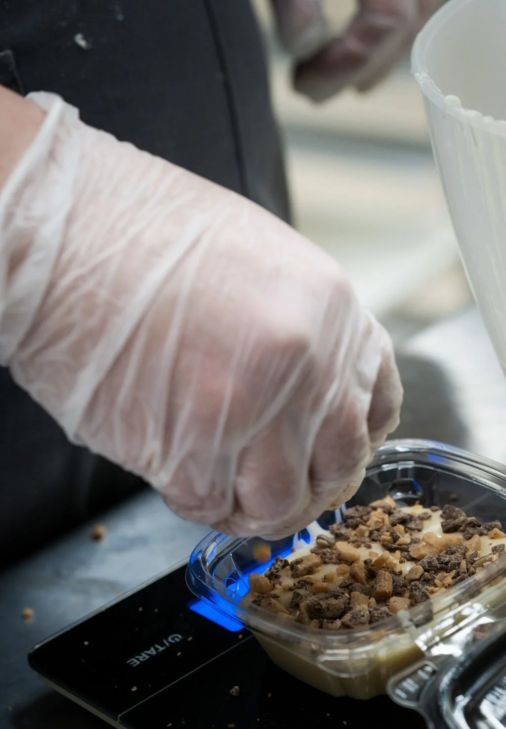 A person wearing a glove pours chopped nuts over a dessert, which is on a digital scale.