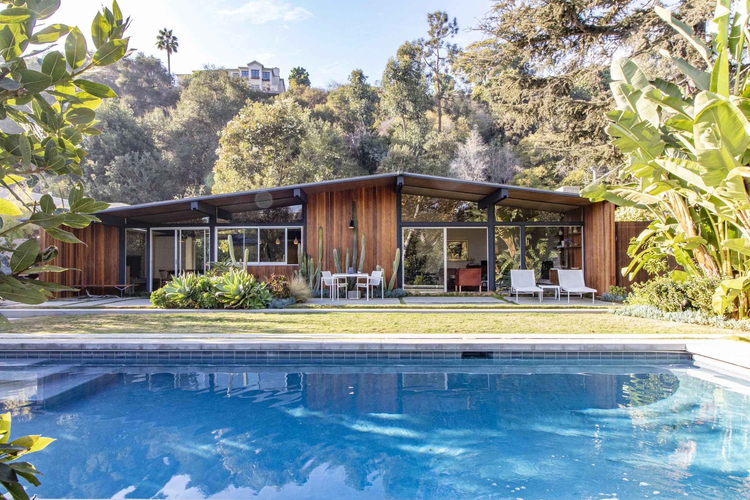 Modern house with wooden exterior, glass walls, and patio, surrounded by lush greenery and a swimming pool in the foreground, hillside with trees and a house in the background.