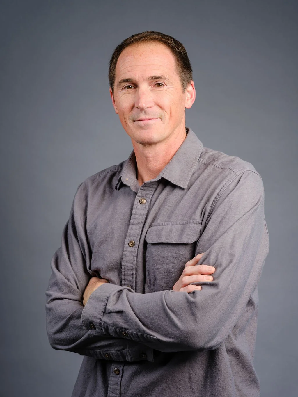Portrait of a middle-aged man with short brown hair, wearing a gray button-up shirt, arms crossed, standing against a plain gray background.