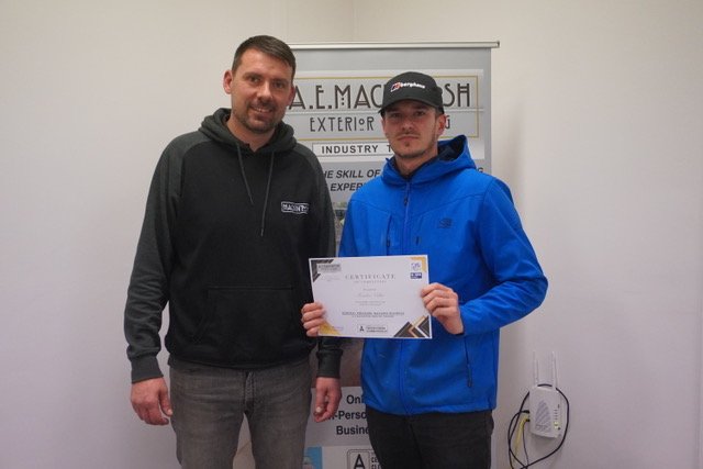 Two men standing next to each other indoors, one holding a certificate, with a banner in the background.