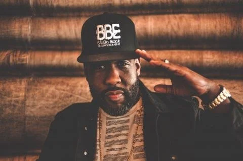 Homme portant une casquette noire avec le logo 'BBC Basic Black', saluant de la main, avec un fond en bois.
