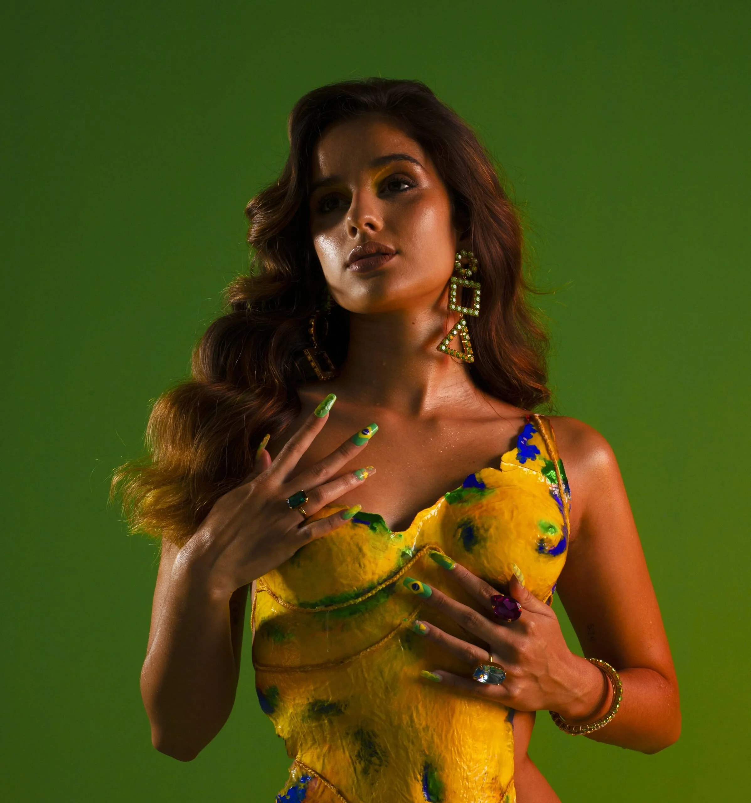 Une femme portant une robe jaune avec des motifs bleus et verts, des boucles d'oreilles géométriques, et des ongles peints aux couleurs du drapeau du Brésil, pose contre un fond vert.
