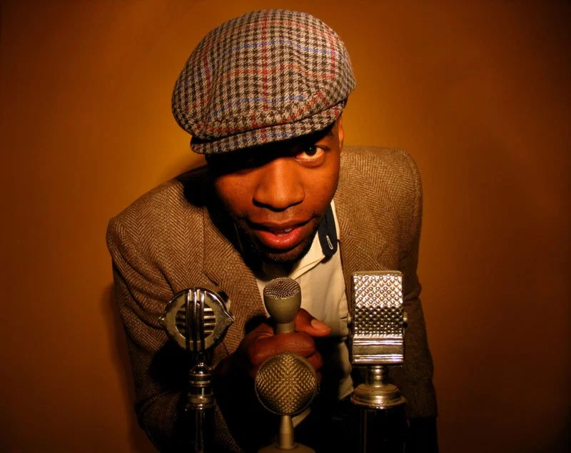 Un homme portant un béret à carreaux, tenant un microphone, entouré de deux autres microphones, regardant l'appareil photo dans une ambiance chaleureuse.