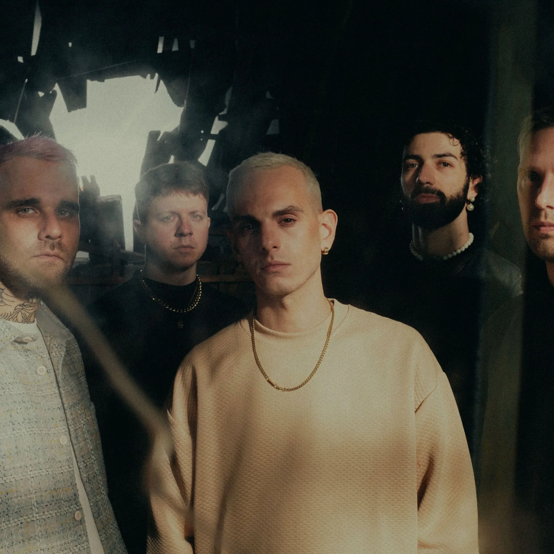 Un groupe de cinq jeunes hommes posant pour une photo, certains portant des bijoux, dans une ambiance sombre avec un éclairage dramatique.