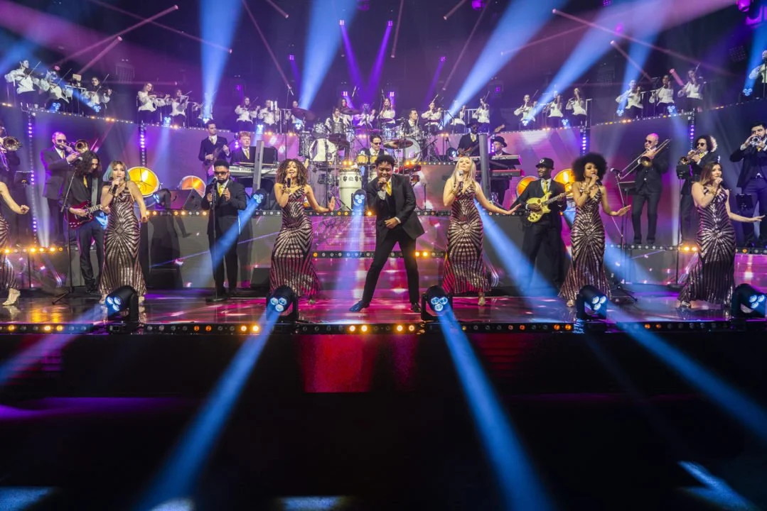Un groupe de musiciens et chanteurs se produisent sur scène avec des éclairages colorés et un orchestre en arrière-plan.