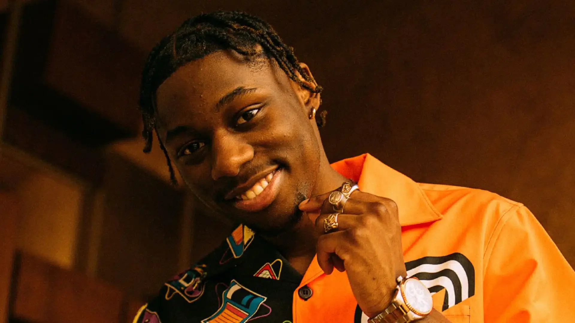Jeune homme souriant portant une chemise orange et des bijoux, avec un fond sombre.
