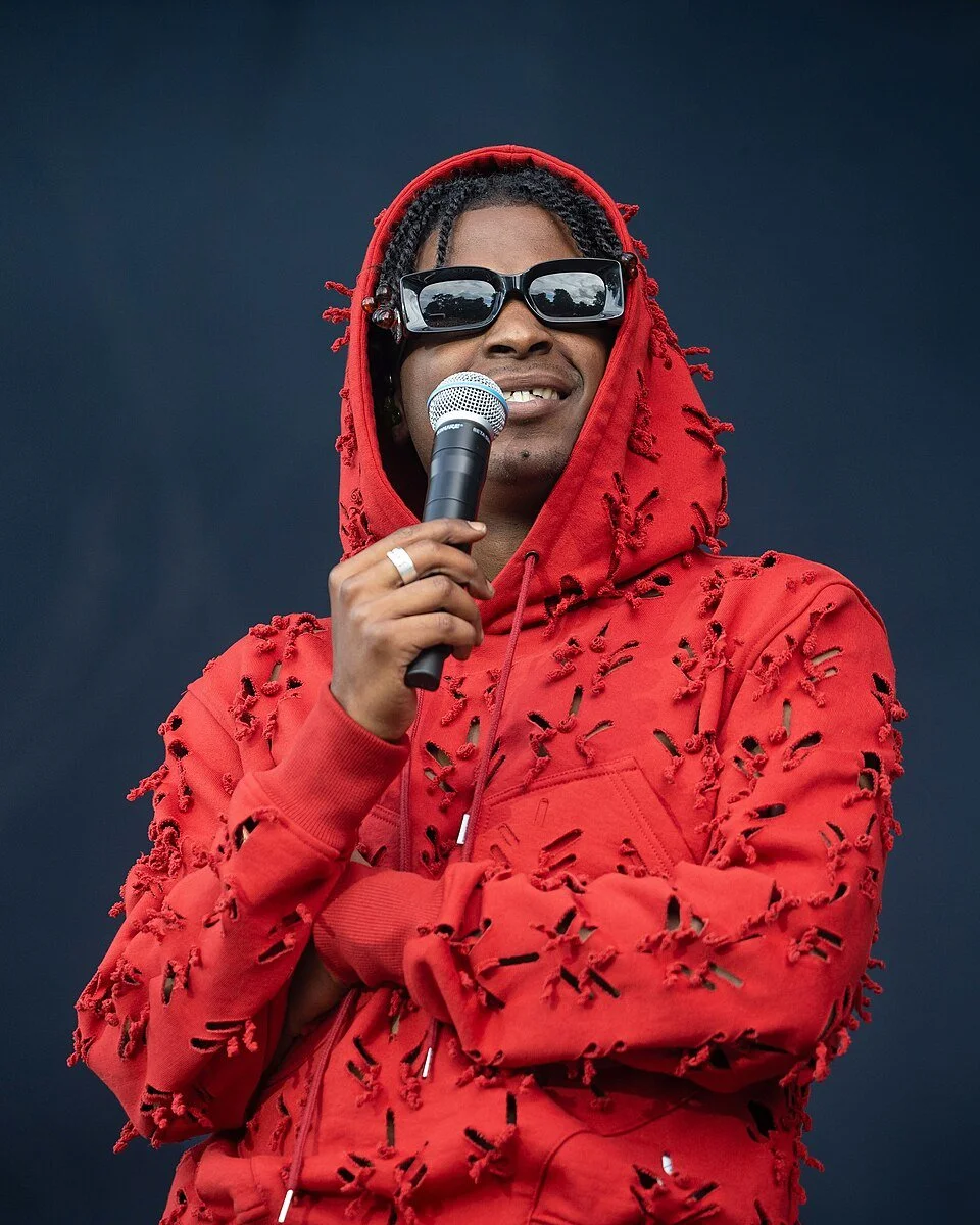 Un homme portant des lunettes de soleil noires, un sweat à capuche rouge déchiré, tient un microphone et sourit lors d'un événement en extérieur.