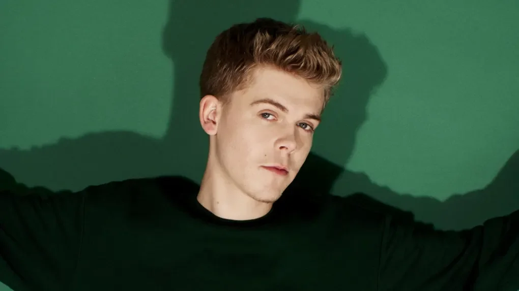 Jeune homme avec cheveux bruns, portant un t-shirt noir, devant un fond vert, regardant vers l'objectif avec une expression sérieuse.