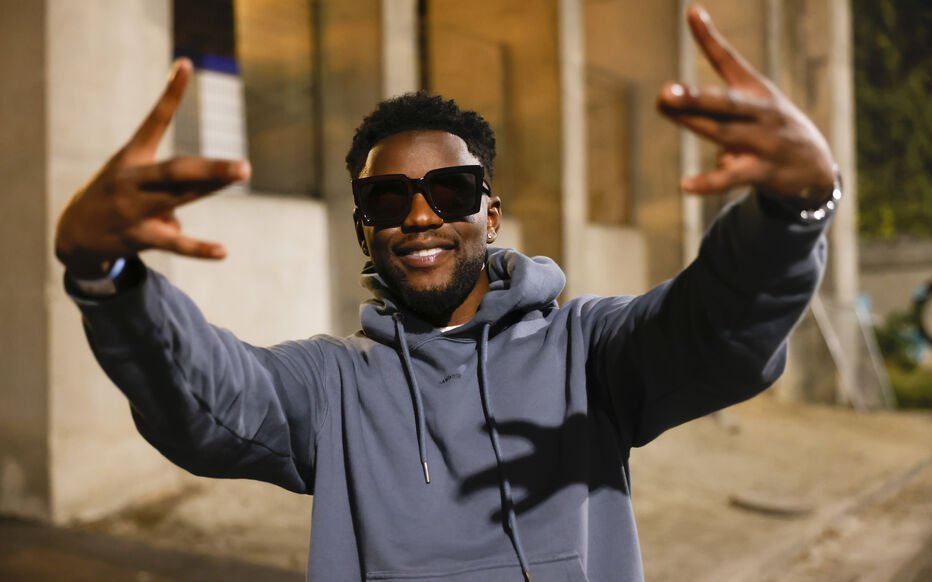 Homme souriant portant des lunettes de soleil et un sweat à capuche, faisant un geste de la main à l'extérieur la nuit.