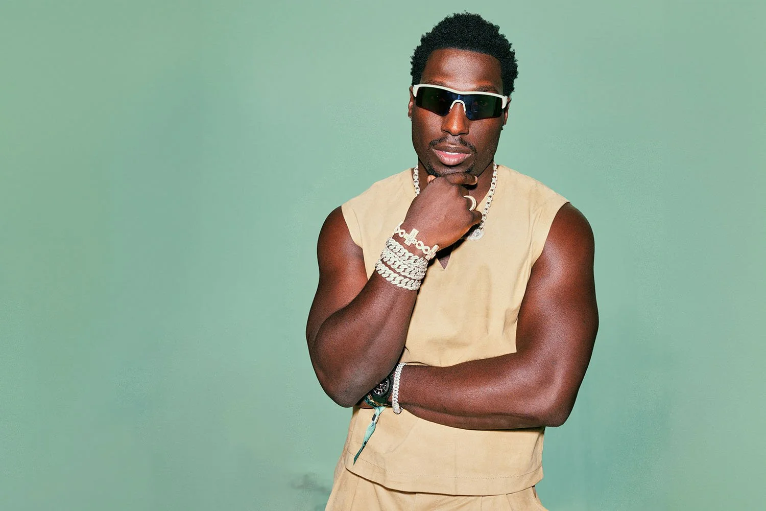 Un homme noir portant des lunettes de soleil noires, un haut sans manches beige, plusieurs bracelets en argent et une chaîne, pose contre un fond vert pastel, arborant une expression confiante.