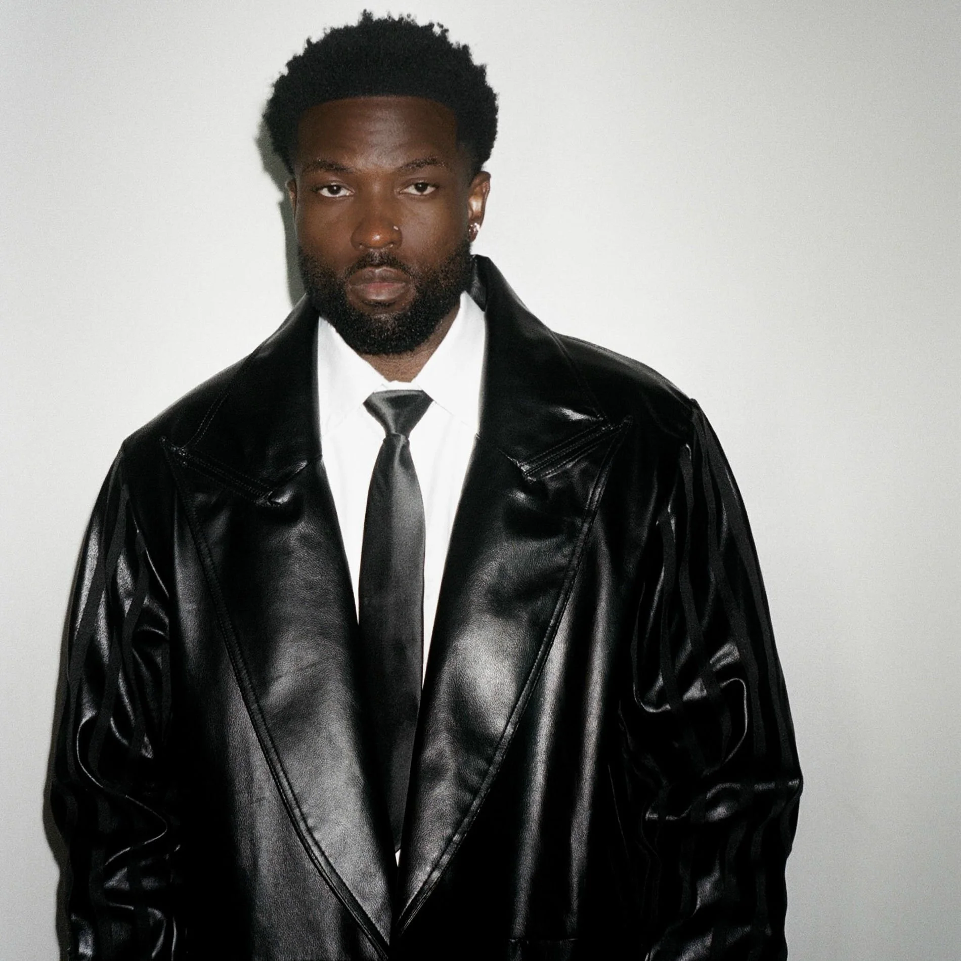 Un homme portant une veste en cuir noir, une chemise blanche, une cravate noire, les cheveux courts et bouclés, avec une barbe, posant devant un mur blanc.