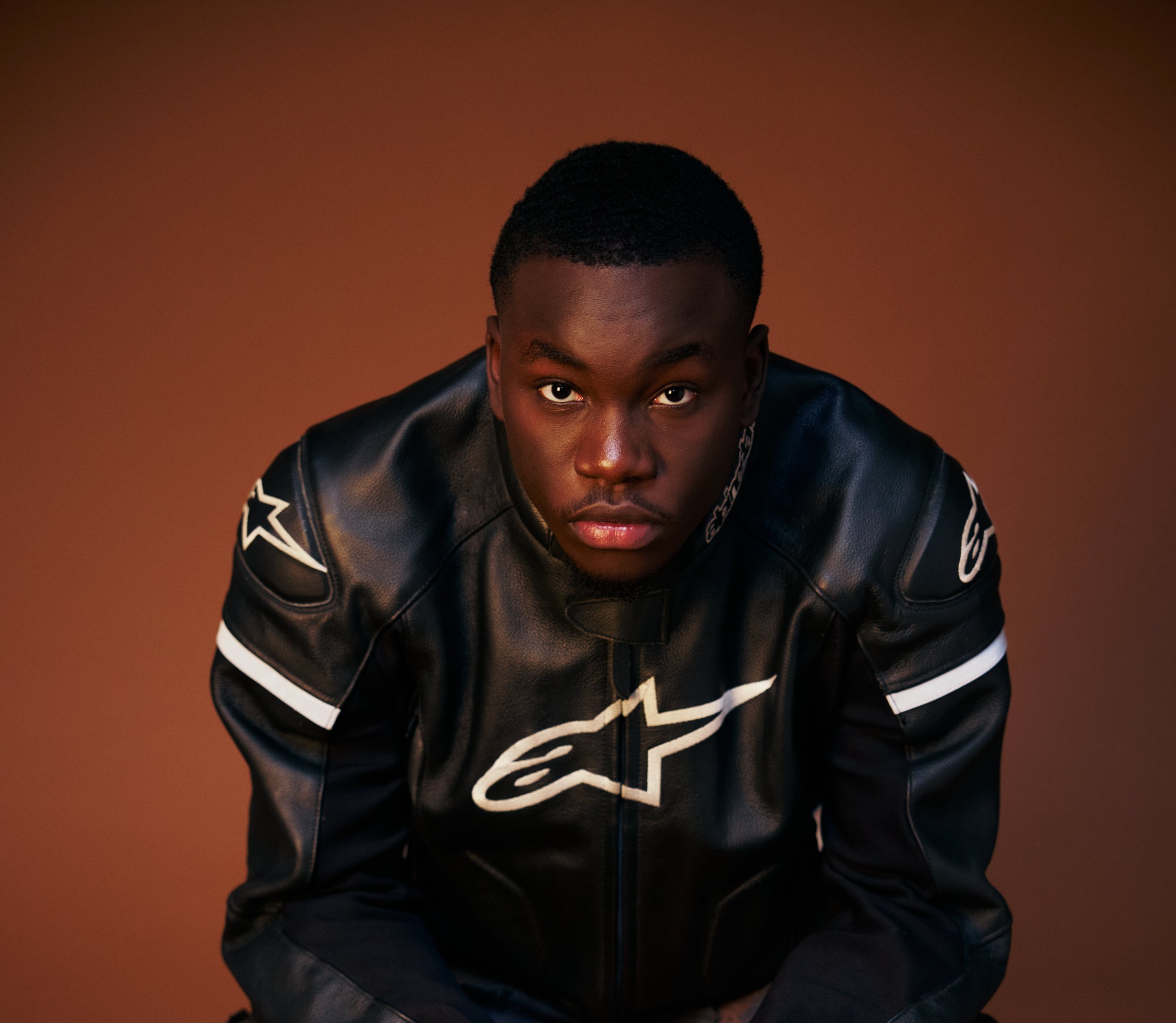 Jeune homme portant une veste en cuir noire avec logo blanc, regardant fixement l'objectif, assis contre un fond marron.