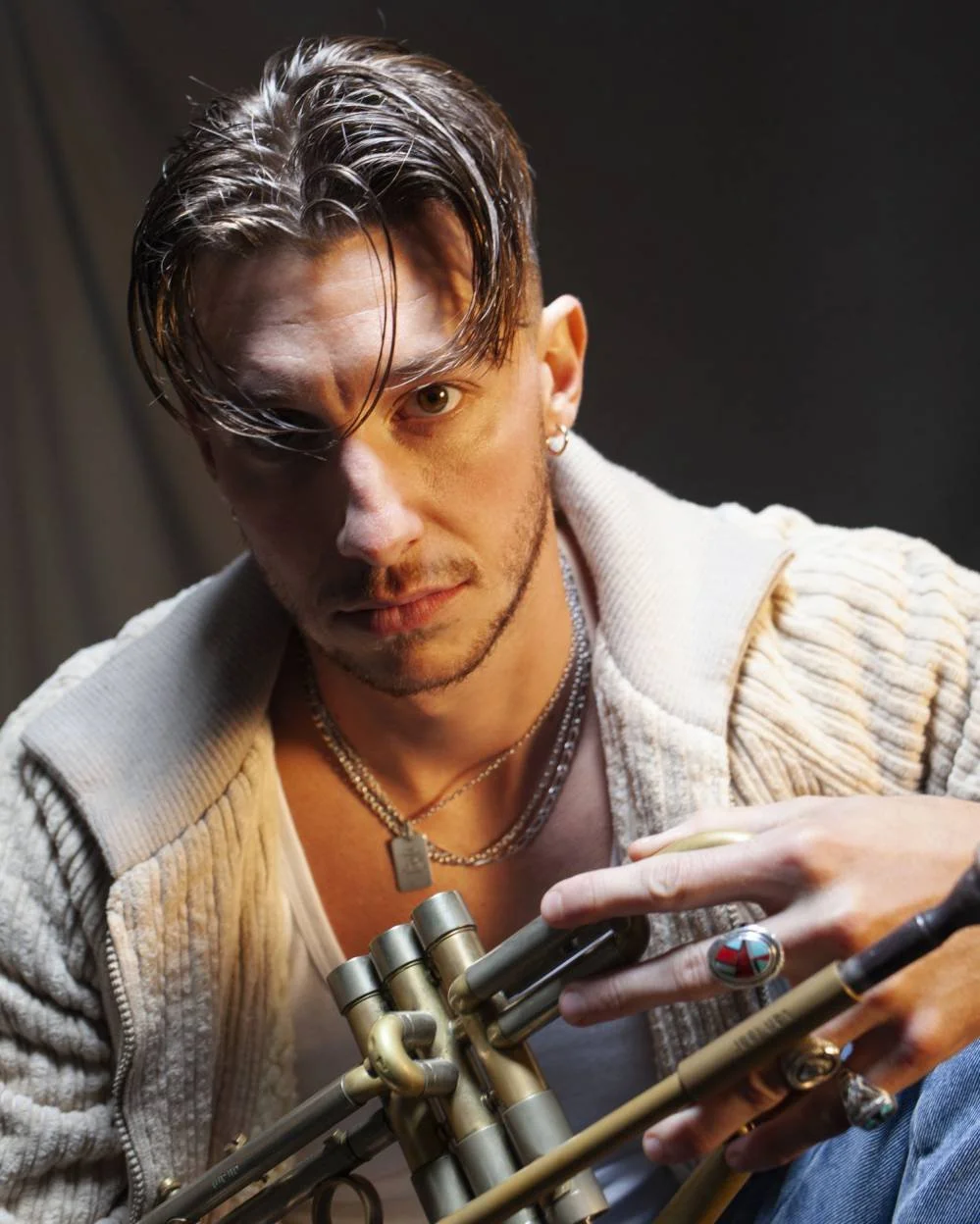 Un homme avec des cheveux foncés et raides, portant plusieurs bijoux, regarde intensément la caméra. Il porte un sweat en laine beige et tient un instrument de musique en métal.