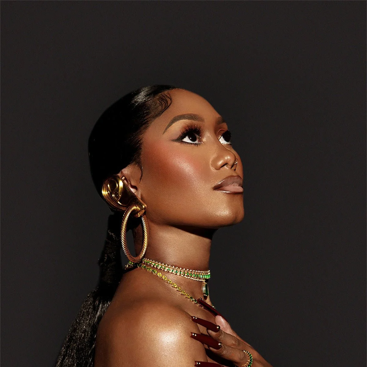 Une femme noire avec un maquillage sophistiqué, portant de grandes boucles d'oreilles en or, un collier chic, et une coiffure lisse, posant contre un fond noir.