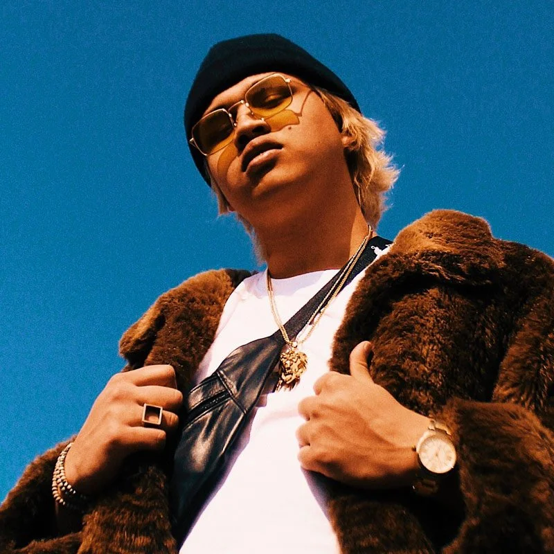 Un homme porte des lunettes de soleil, un bonnet noir, une veste en fourrure marron et une montre, posant contre un ciel bleu.