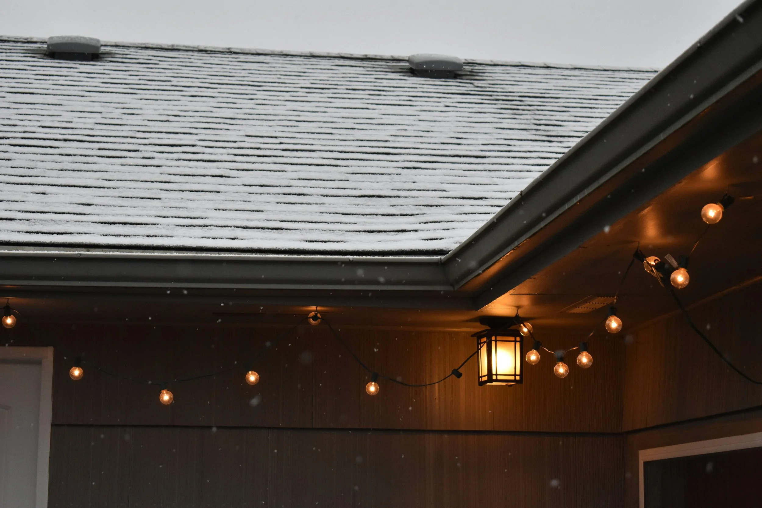 Part of a house with snow on the roof, snowflakes falling, and warm string lights and a lantern-style porch light on the exterior wall.