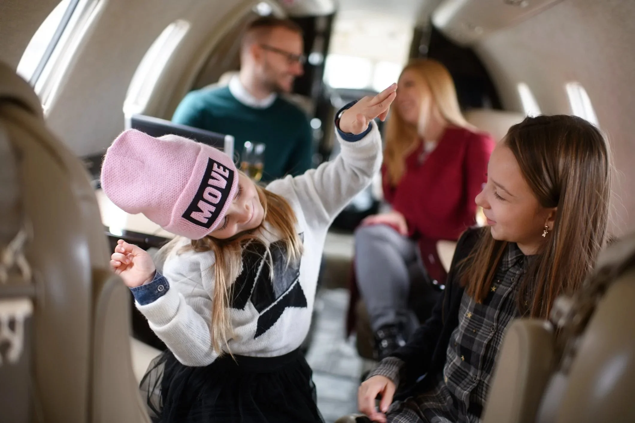 A young girl with a pink beanie and white sweater dancing and smiling inside an airplane cabin, with other passengers in the background.