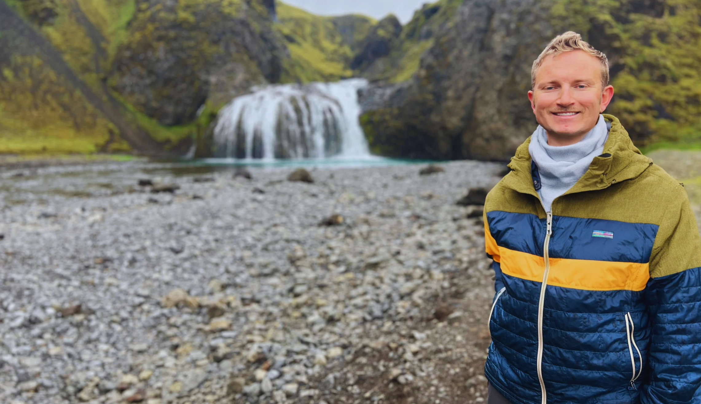 A man smiling outdoors near a rocky riverbank with a waterfall and green hills in the background.