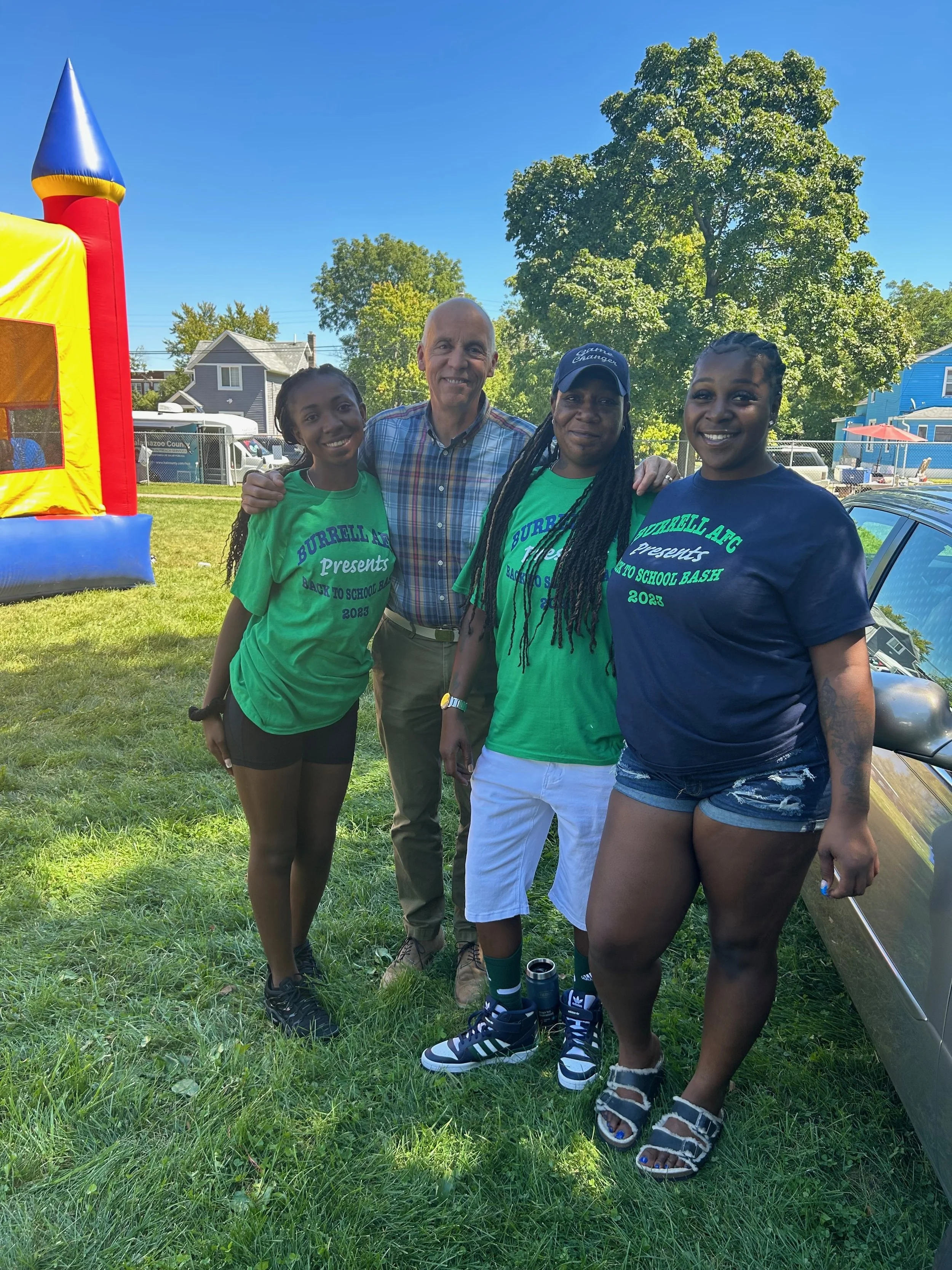 Four people, three women and one man, are outdoors at a park event on a sunny day. Two women are wearing green shirts that say 'Burrell AFG Presents Back to School Bash 2023.' They are standing next to a man in a checkered shirt and beige pants, and a woman in a navy shirt and denim shorts. They are smiling, standing on grass, with a bouncy house and houses in the background.