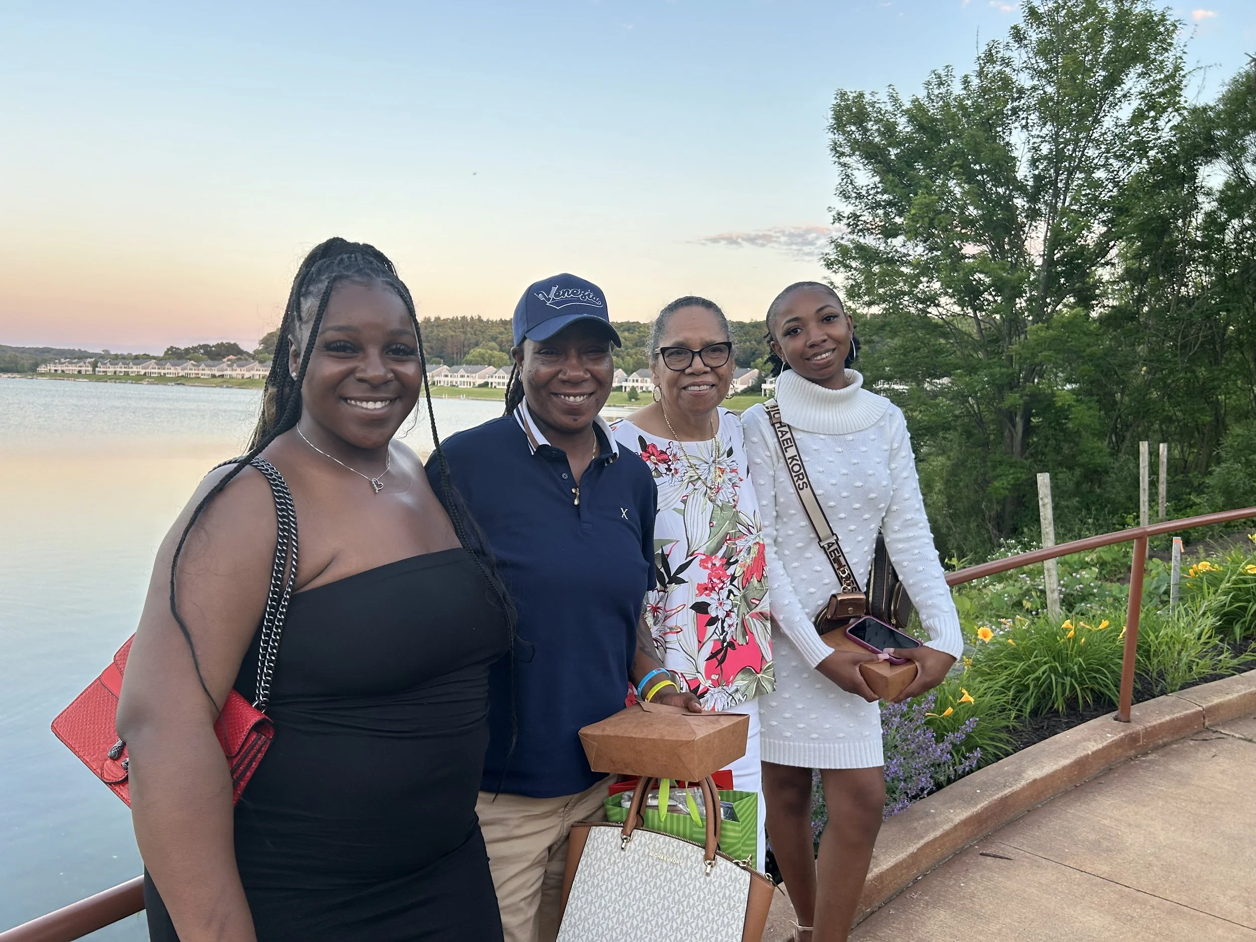 Four women standing outdoors near a water body with trees and houses in the background, smiling for the camera during sunset.
