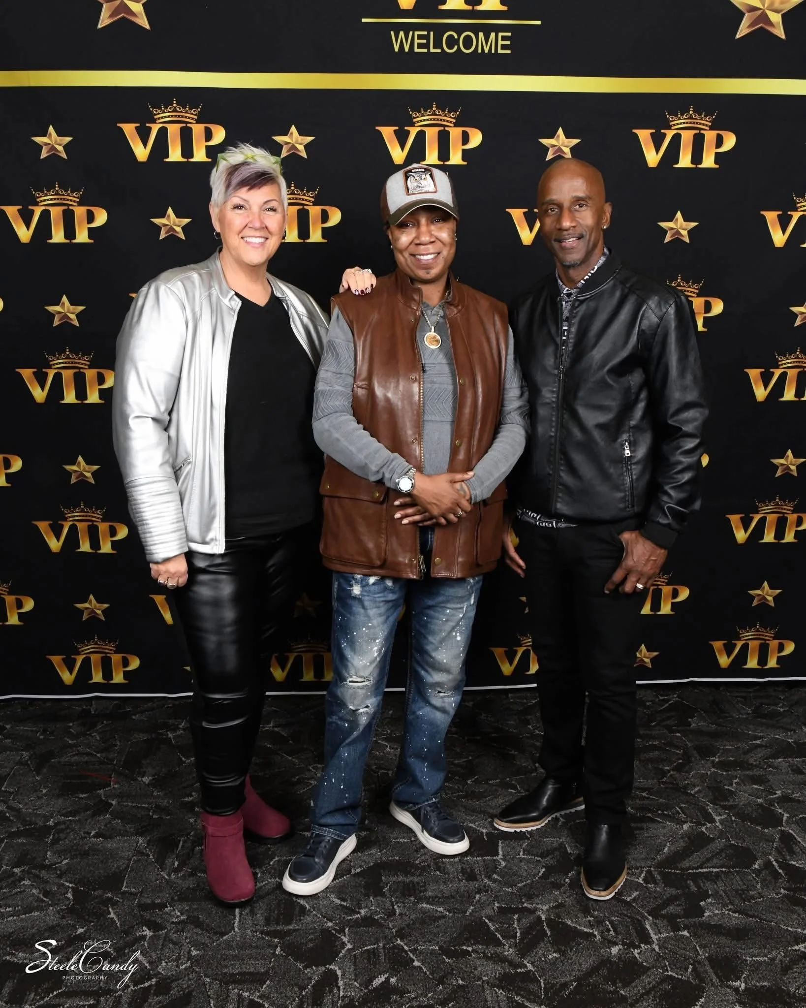 Three people standing in front of a backdrop with gold stars, crowns, and 'VIP' and 'WELCOME' text, smiling at the camera.