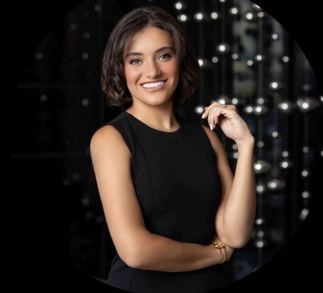 A young woman with short, wavy brown hair and light makeup, wearing a sleeveless black dress, smiling confidently and standing with her arm crossed and her hand near her face, in front of a dark background with decorative lights.