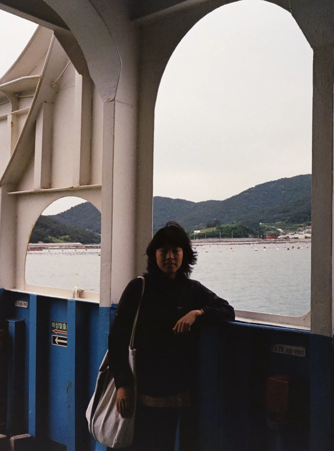 Persona en la cubierta de un barco mirando hacia la cámara, con paisaje de montaña y agua al fondo.