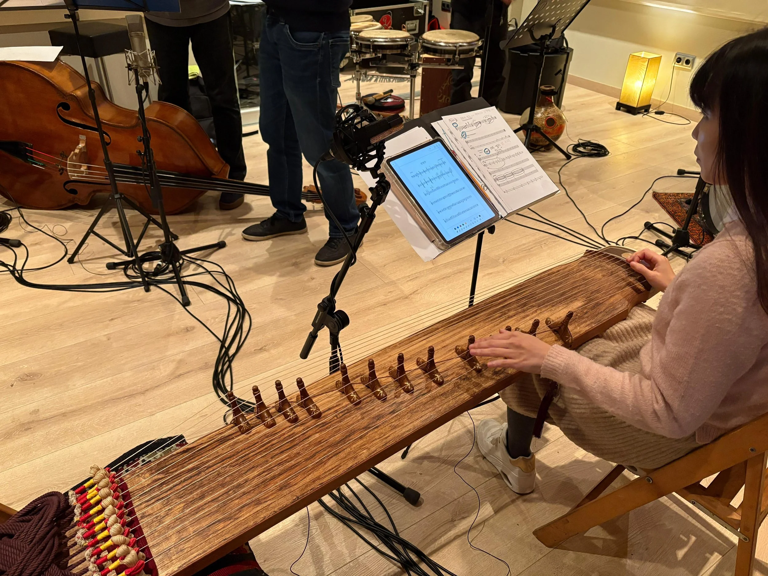 Una joven tocando un instrumento de cuerda tradicional, sentado en una silla, con partituras y un tablet delante. En el fondo, otros músicos y instrumentos musicales.
