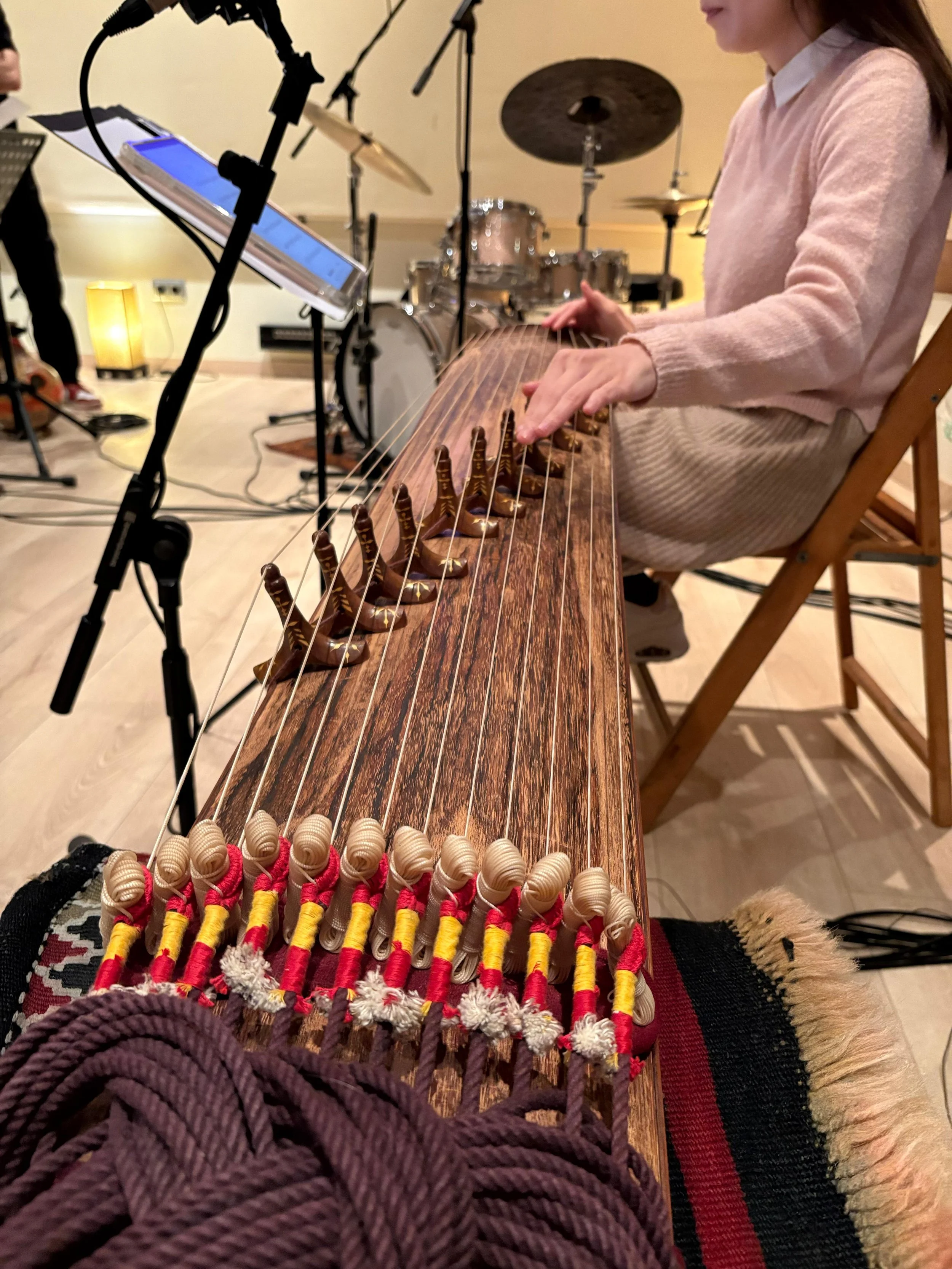 Mujer tocando zither en un estudio, con instrumentos musicales y micrófonos en el fondo.