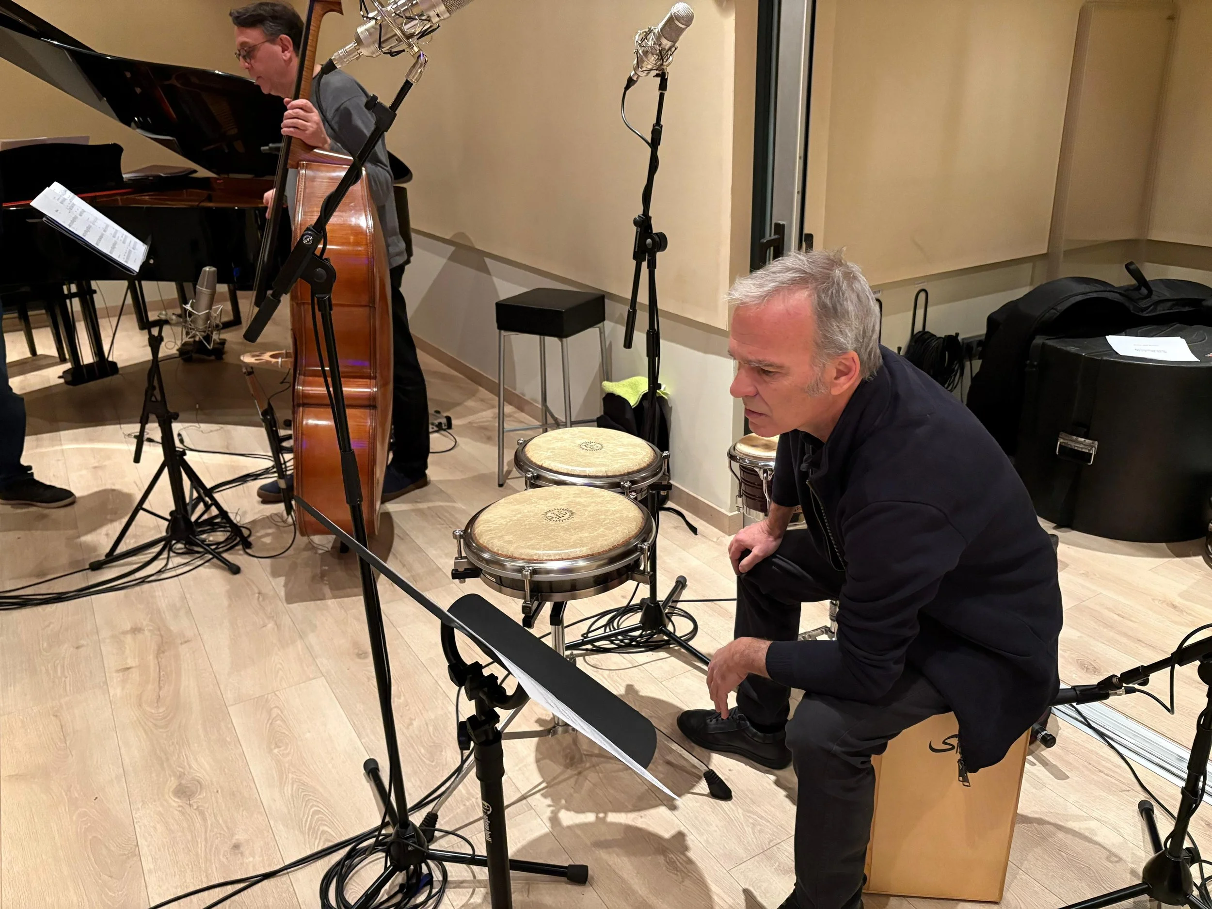 Hombre con cabello gris sentado en un banco, observando atentamente en un estudio de grabación, con instrumentos musicales y micrófonos alrededor.