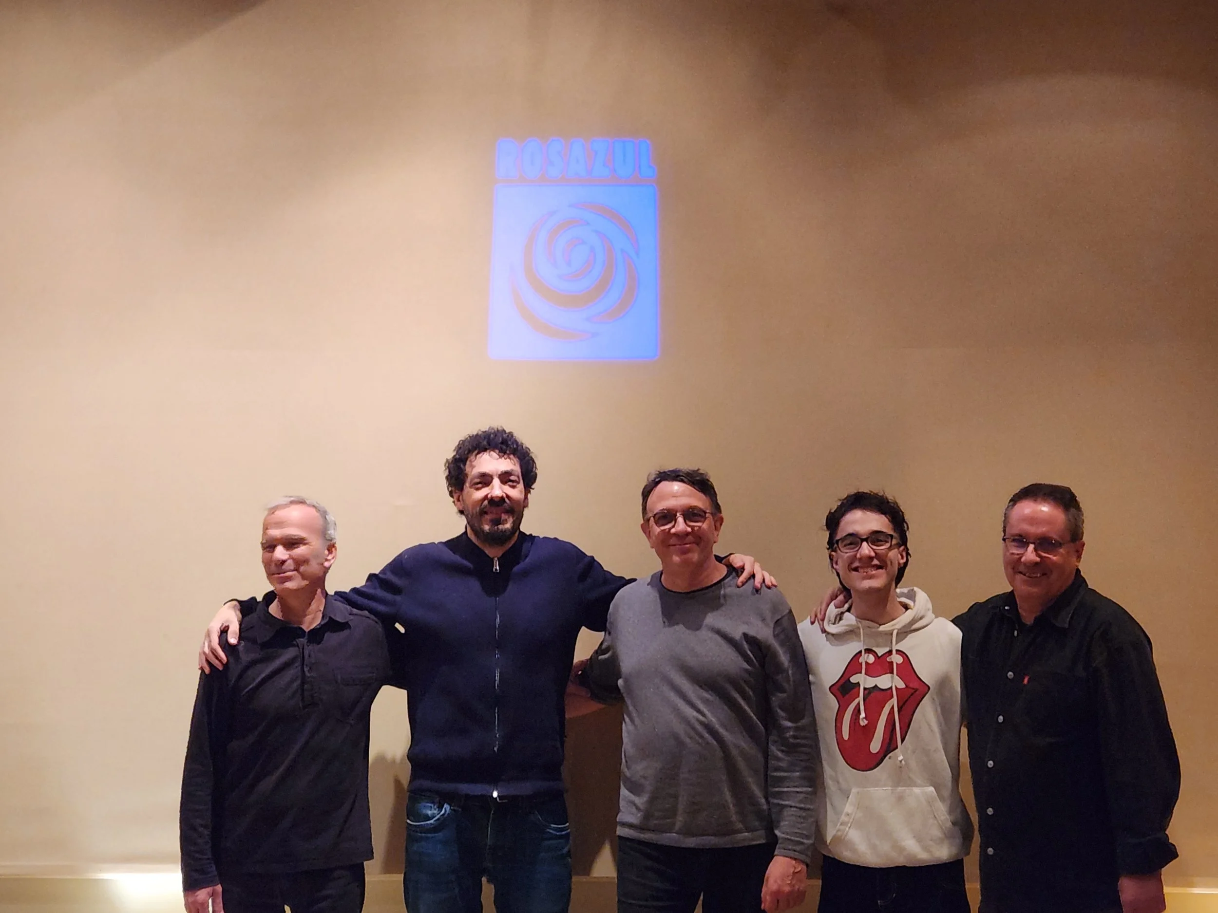 Cinco hombres de pie en una fila, sonriendo y con los brazos sobre los hombros de las personas a su lado, en un fondo de pared beige con un logo que dice "ROSAL" y tiene un diseño de espiral azul.