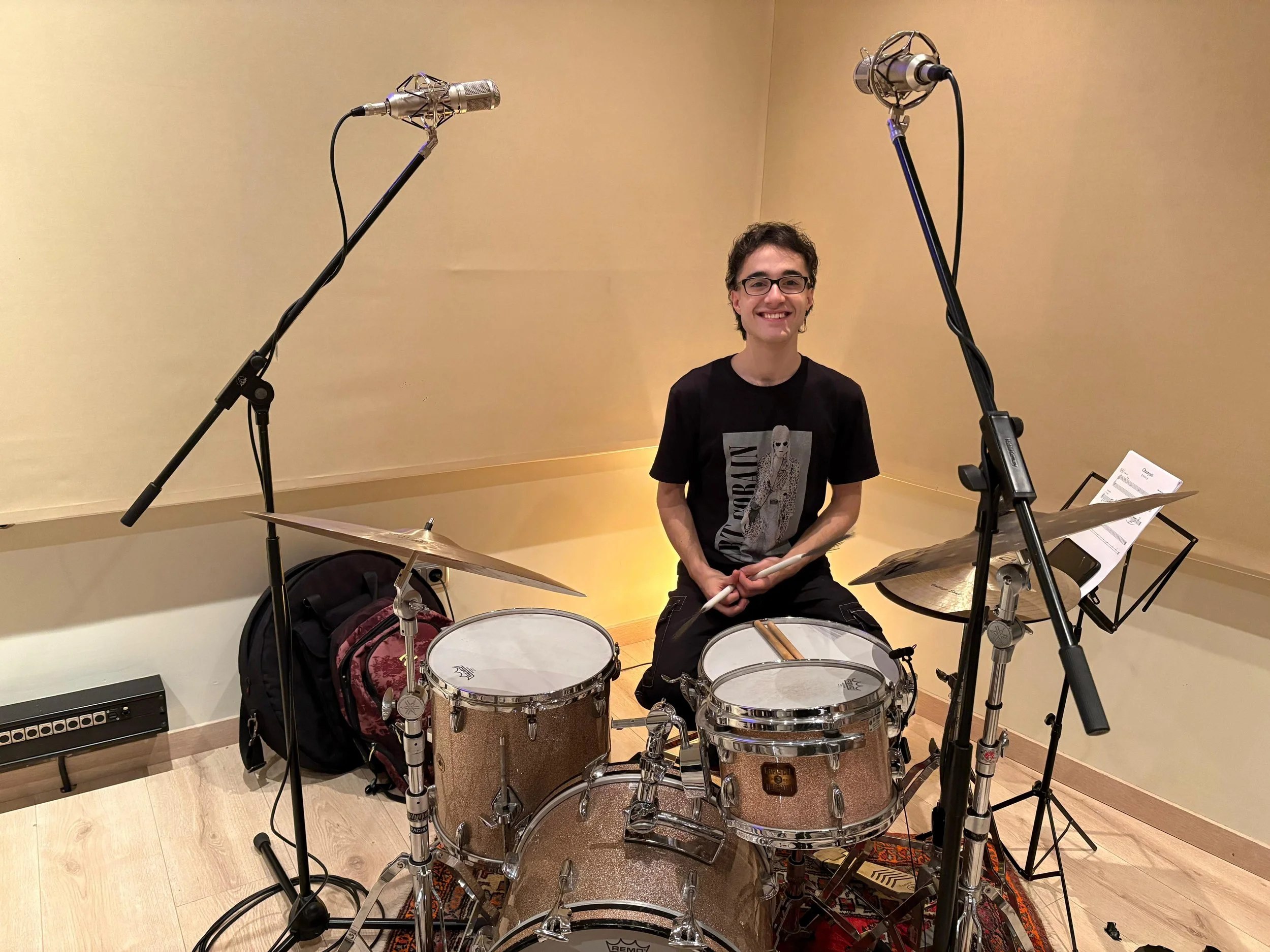 Un joven sentado tras bateria en estudio de grabación, con micrófonos y partituras alrededor.