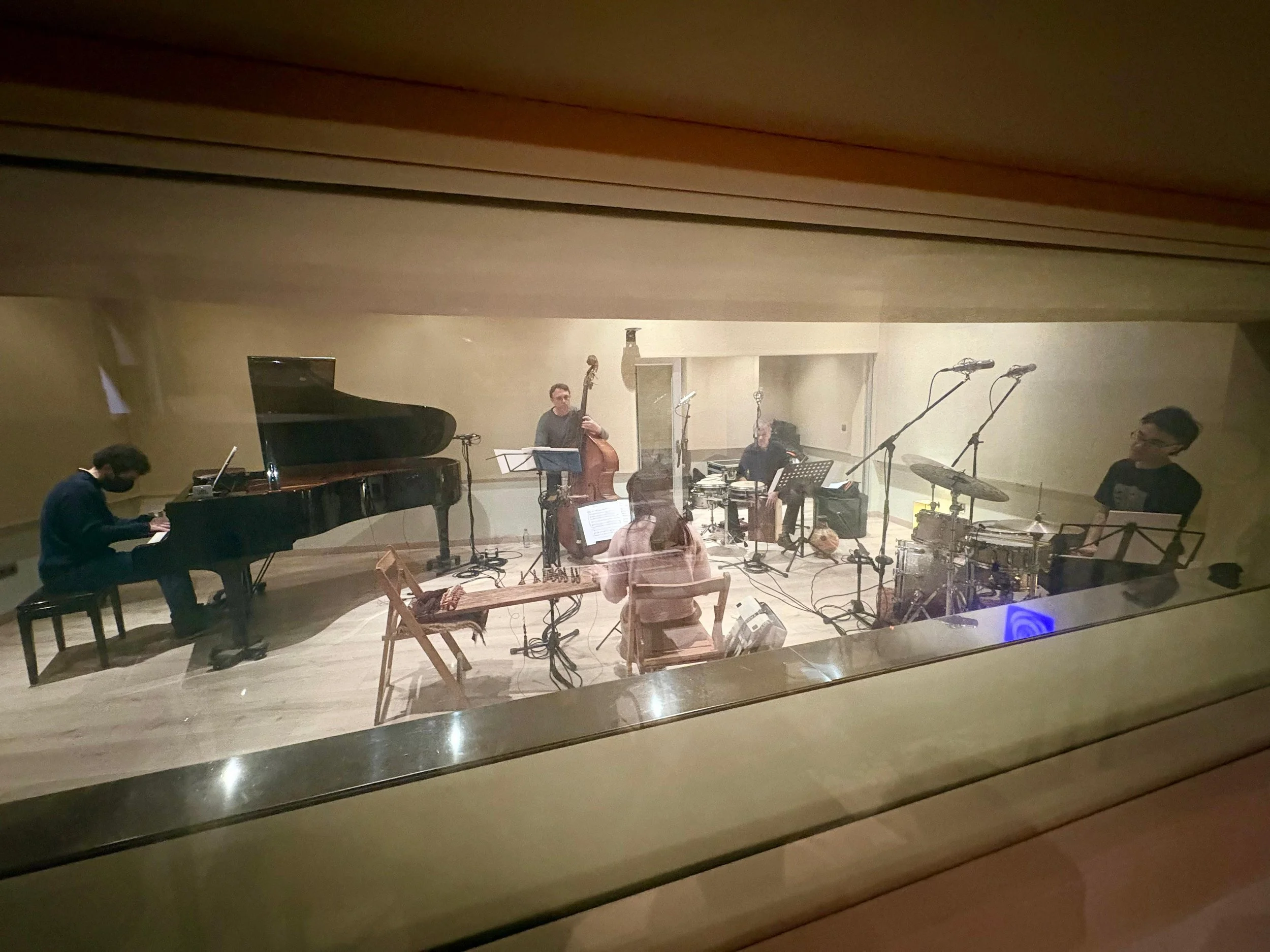 Cuatro músicos tocando instrumentos en un estudio de grabación, vista a través de una ventana de vidrio.