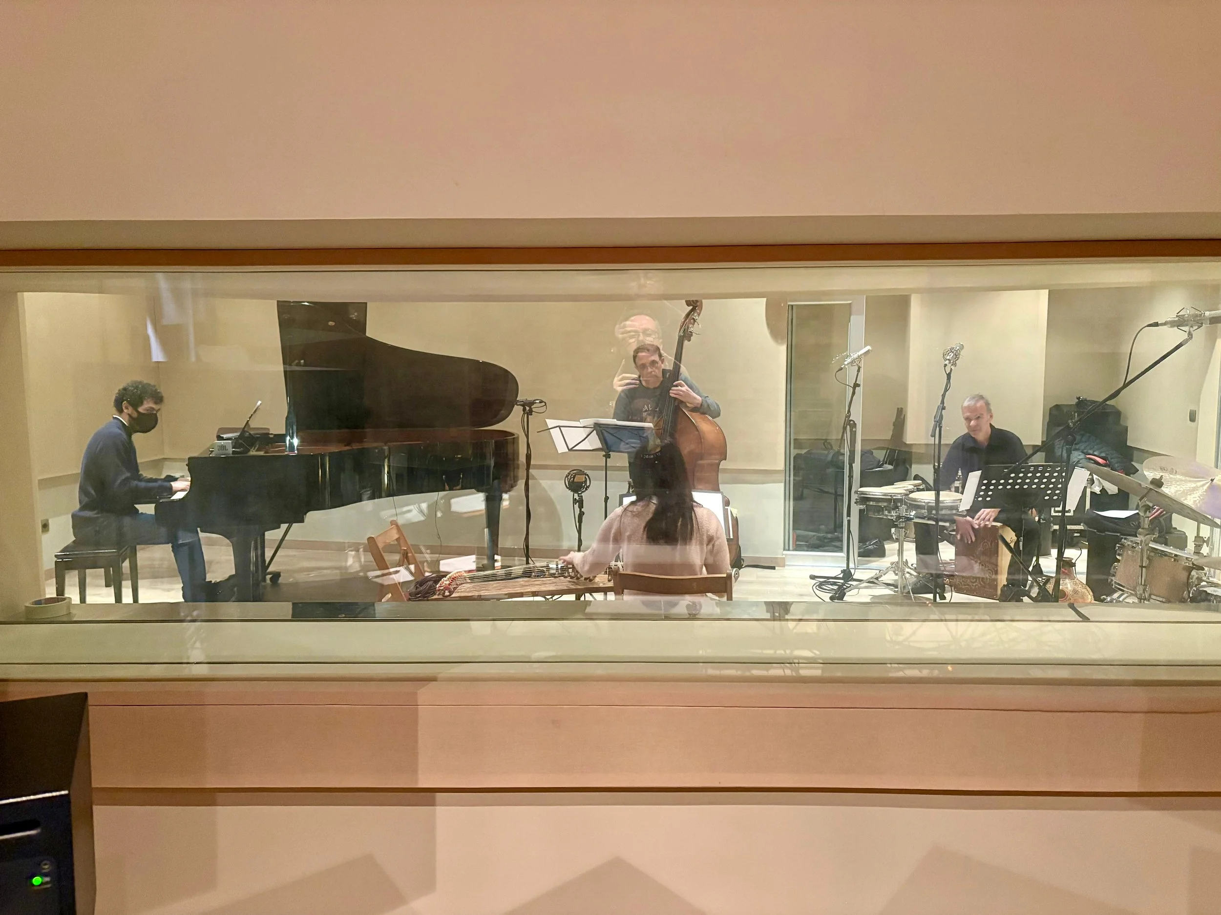 Cuatro músicos tocando instrumentos en un estudio de grabación. Un pianista, un bajista, un percussionista y una mujer sentada con un piano de cola.
