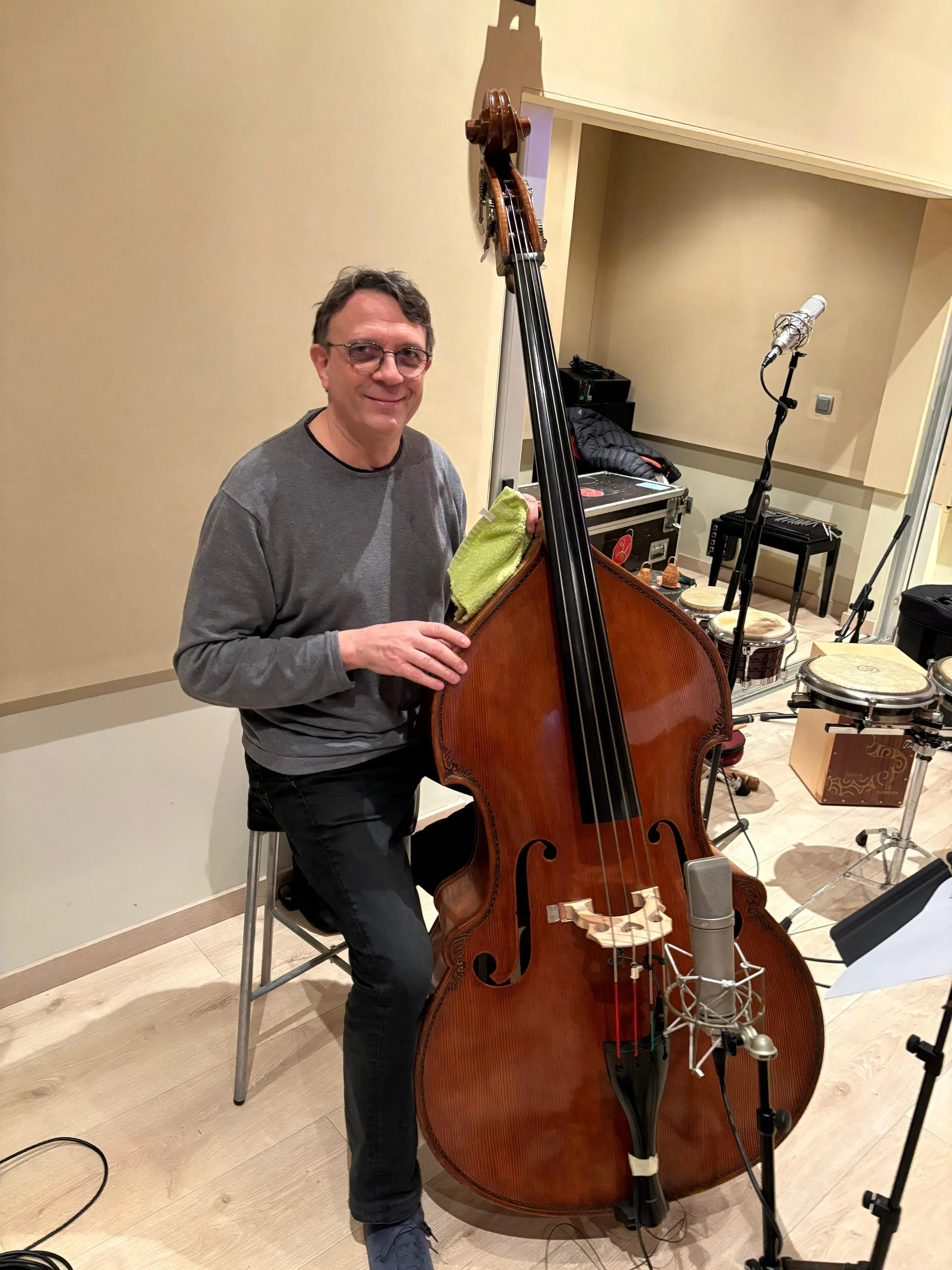 Hombre sentado en un estudio de grabación, sosteniendo un contrabajo, con equipo de grabación y percusión en el fondo.