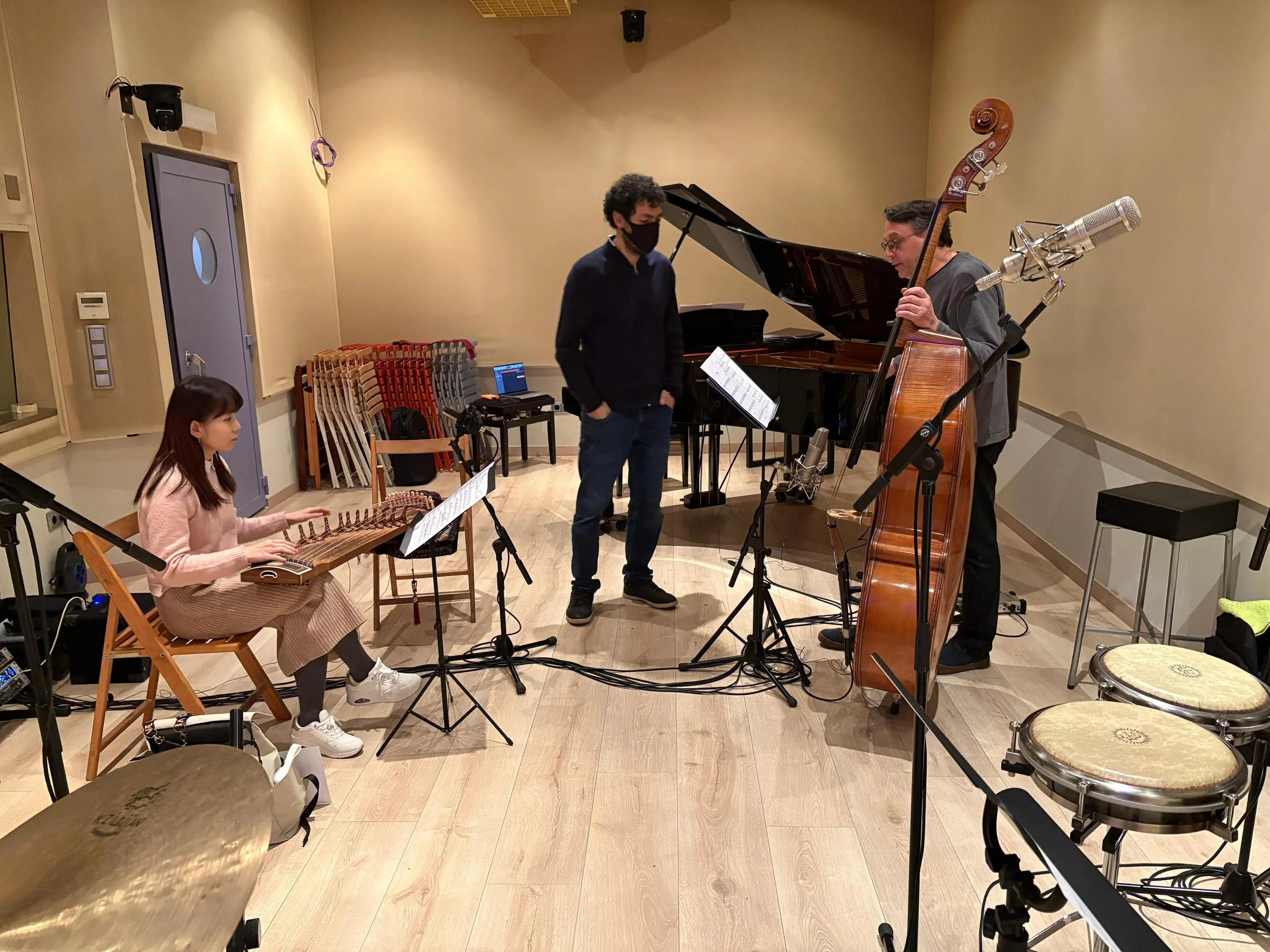 Tres músicos en un estudio de grabación: una mujer tocando un arpa, un hombre de pie y vestido con gafas, y otro hombre tocando un contrabajo. En el fondo hay un piano de cola y diversos instrumentos y equipos de grabación.