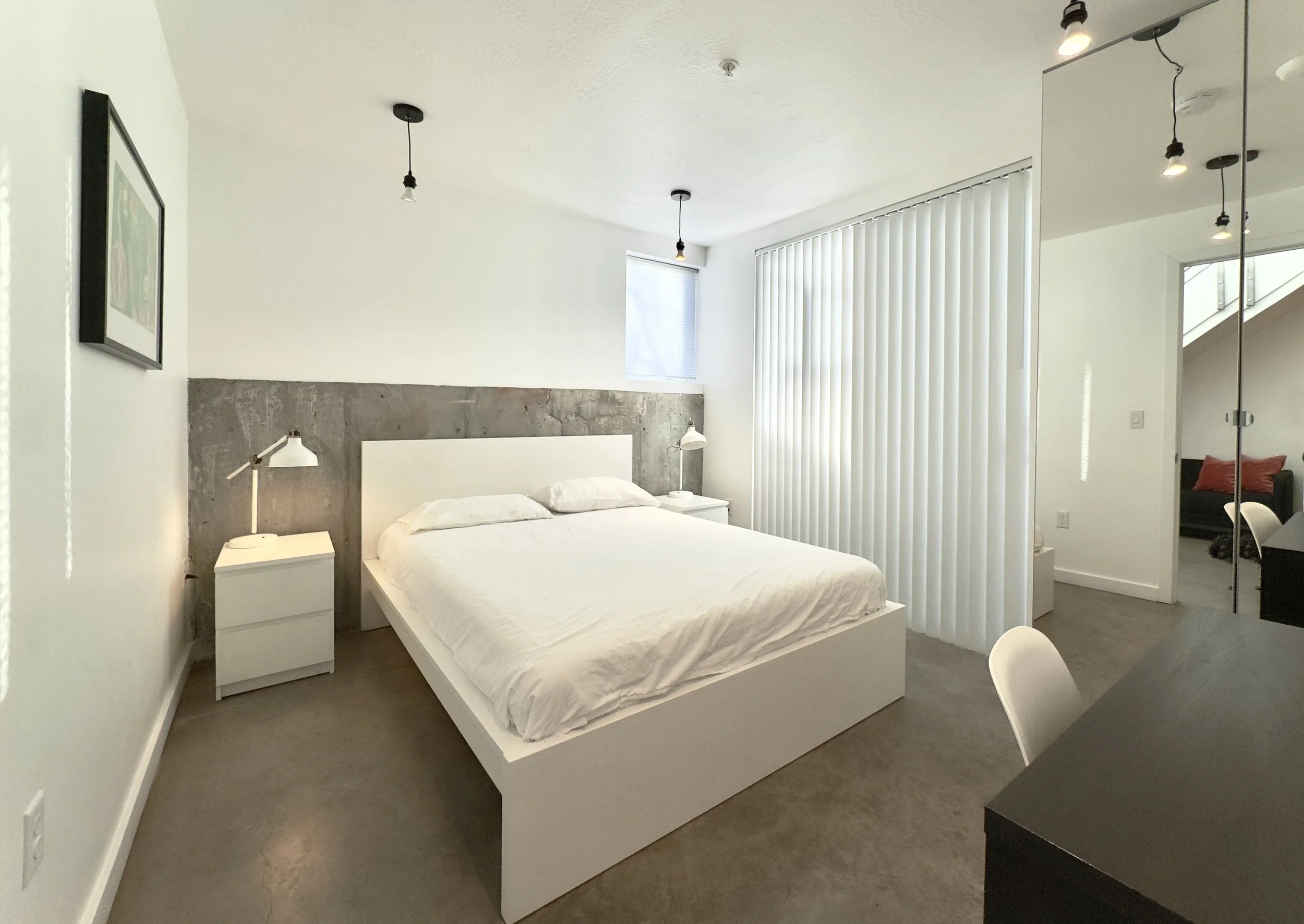 Minimalist bedroom with white bed, two nightstands with white lamps, concrete accent wall, window with blinds, mirror wardrobe, and adjacent room visible through an open doorway.