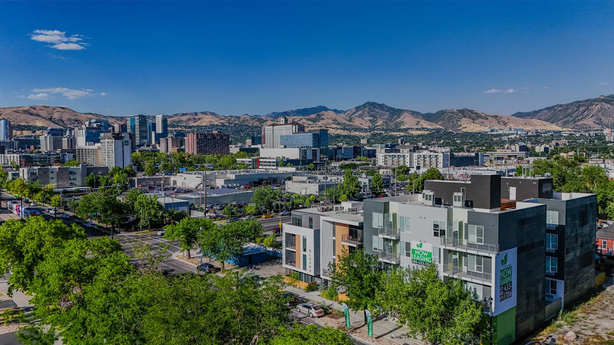A cityscape with mid-rise buildings, parking lots, and green trees, set against a backdrop of mountains on a clear day with blue sky.