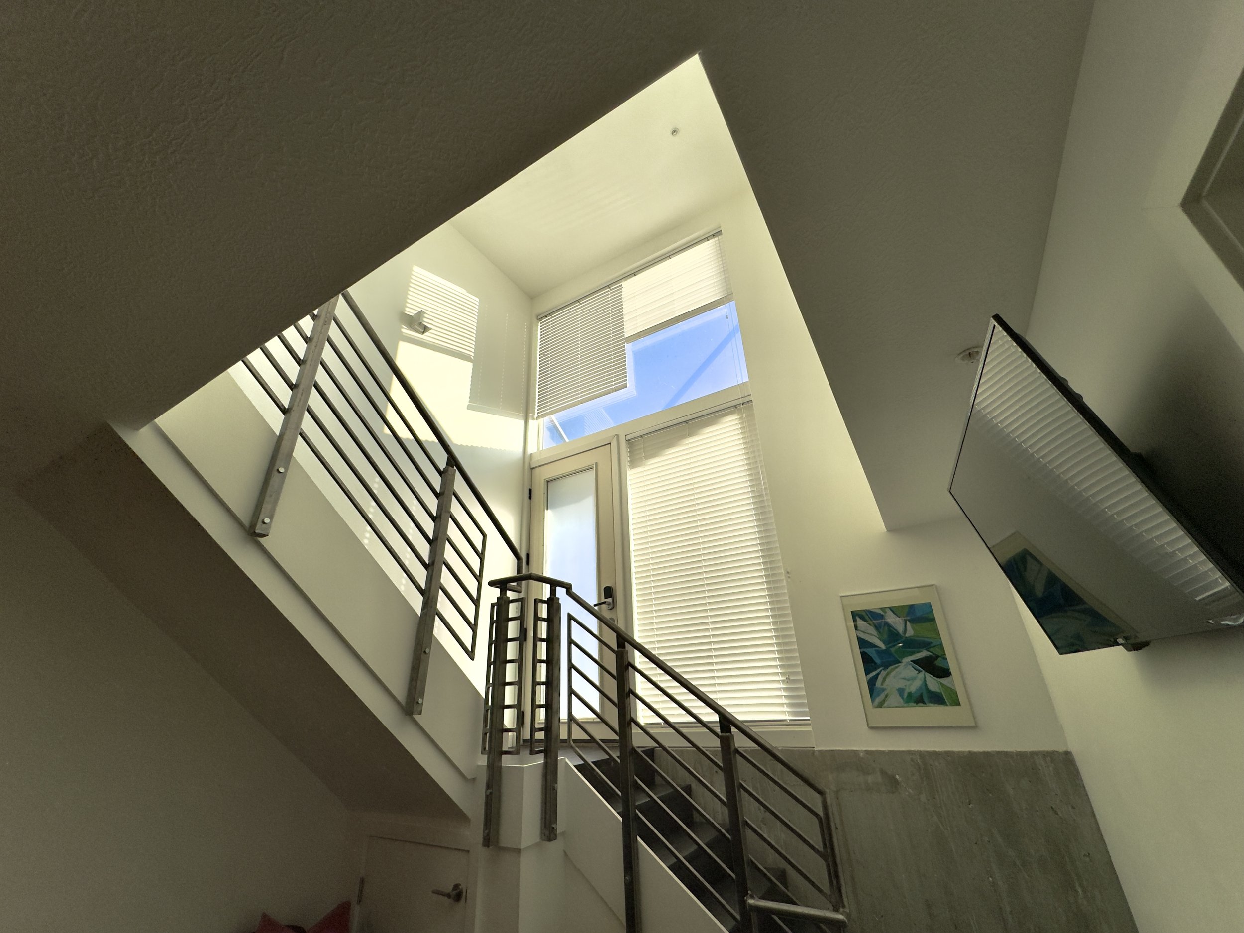 Interior view of a modern staircase with metal railings leading to a bright upper level with large windows and blinds, and artwork on the wall.