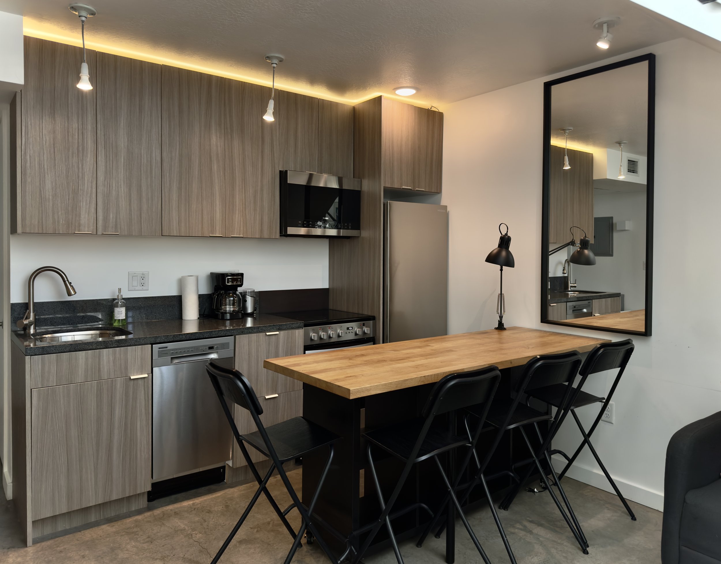 Modern kitchen with wood cabinets, black countertop, stainless steel appliances, and a wooden dining table with black chairs. There is a large mirror on the wall reflecting part of the kitchen and a black table lamp on the table.