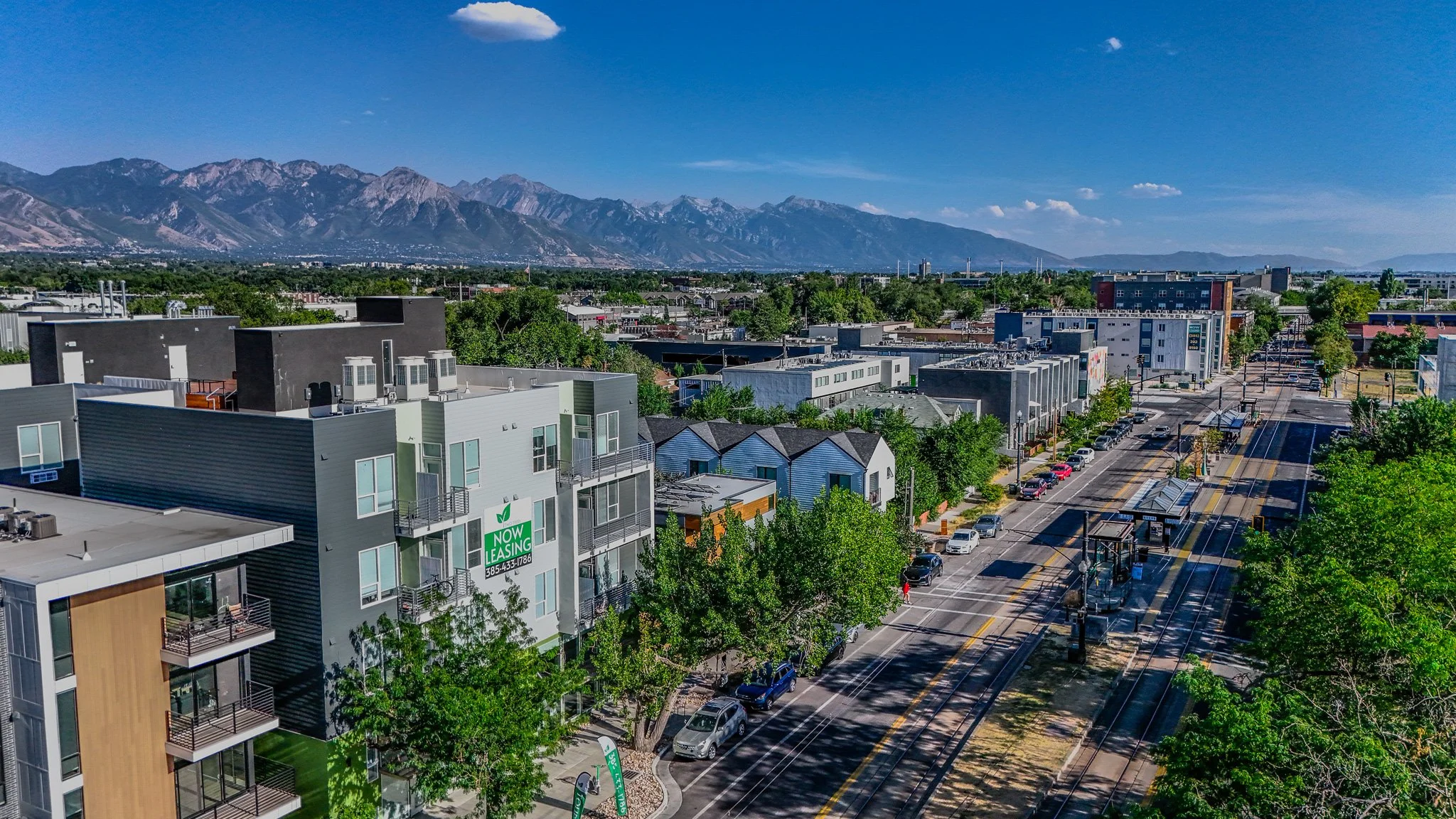 A cityscape view of a street with parked cars, residential buildings, and trees, with mountains in the background under a partly cloudy sky.