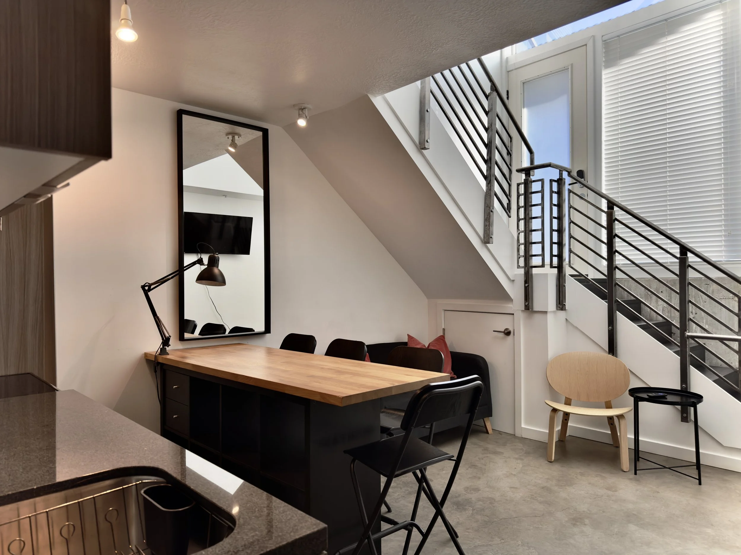 Interior of a modern apartment with a dining area, black chairs, a wooden table, a large mirror on the wall, a flat-screen TV, a black sofa with pink cushions, a wooden chair, and large windows with blinds next to a staircase.