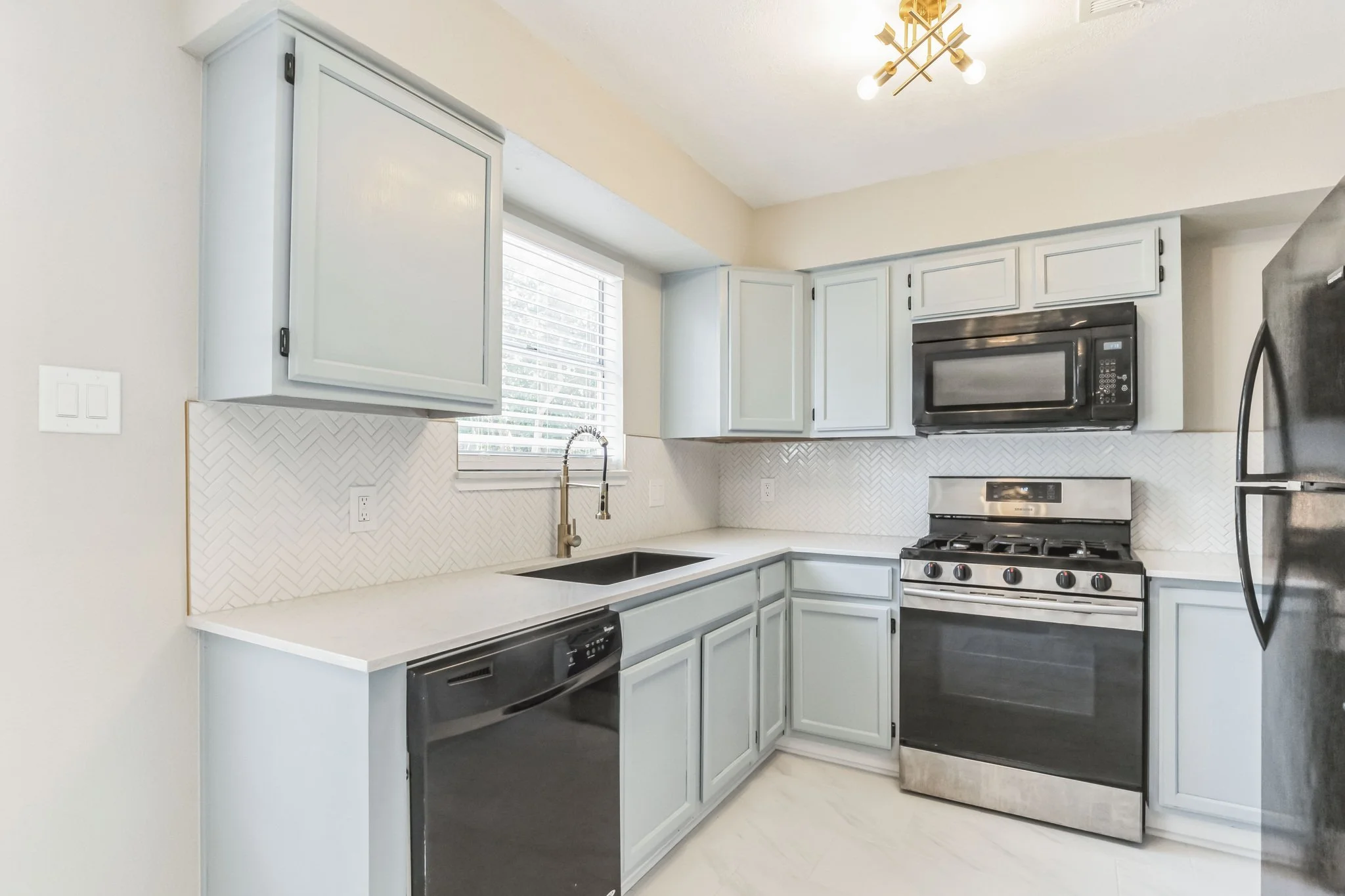 Modern kitchen with light gray cabinets, a black dishwasher, a gas stove, a black microwave, a refrigerator, a window with blinds, and beige walls.