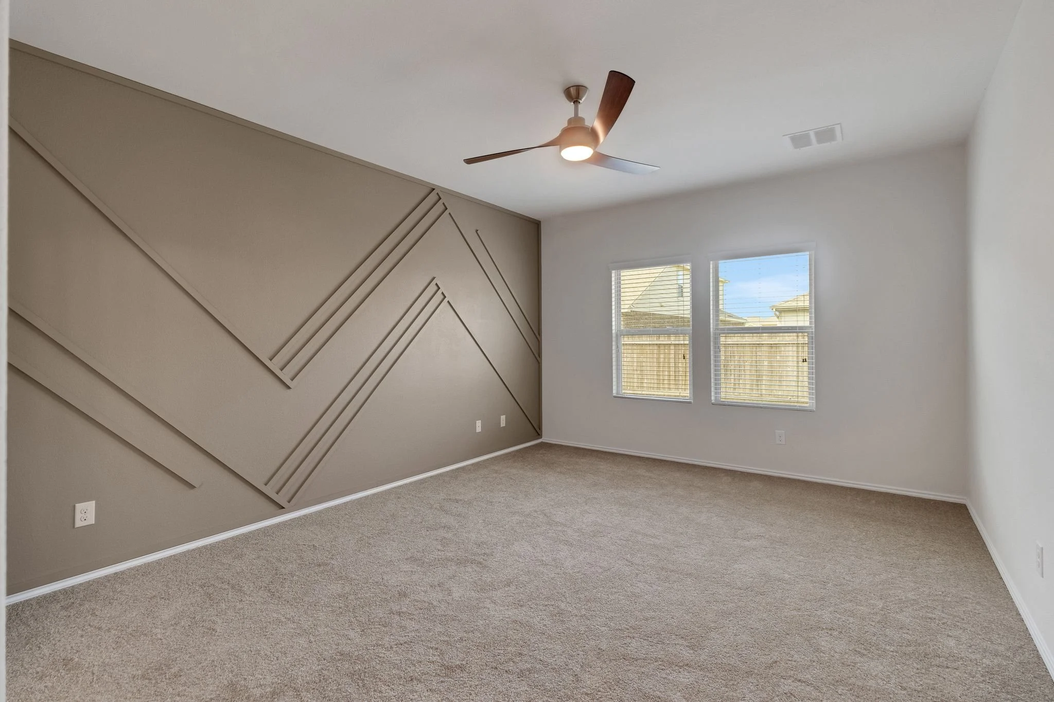 Empty bedroom with beige carpet, white walls, a ceiling fan with wooden blades, and two windows showing a wooden fence outside.