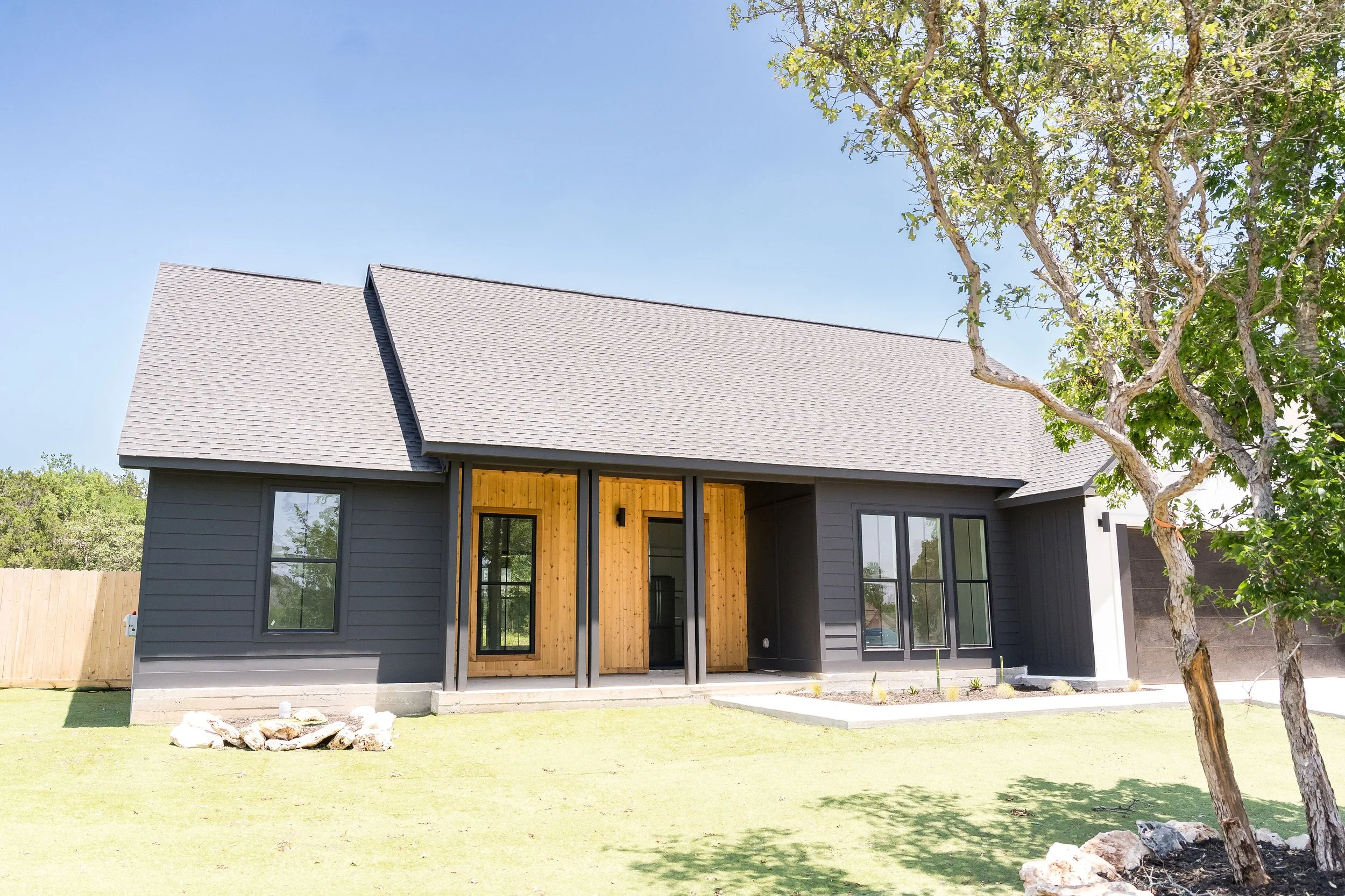 Modern house with dark exterior siding, large black-framed windows, wooden accents at the entrance, a small yard with a tree on the right, and a clear blue sky.