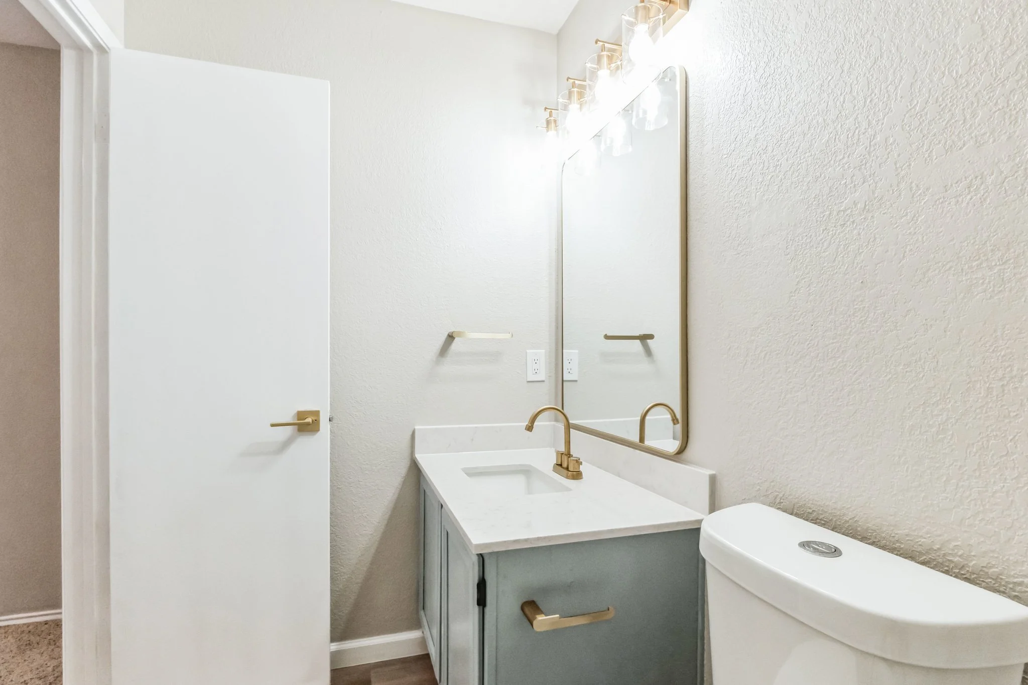 Bathroom with a white door, a green vanity with a marble top, a gold faucet, a large wall mirror, a wall-mounted towel bar, and a white toilet.