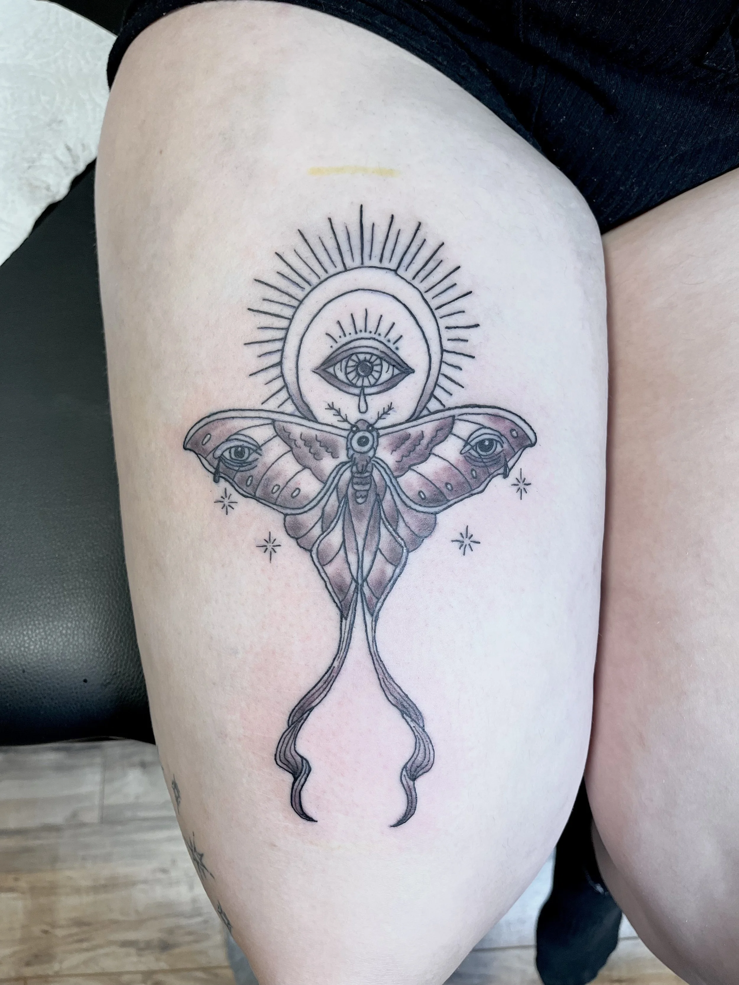 Tattoo of an eye within a halo above a moth with elongated, flowing antennae and wings decorated with eyes, surrounded by star-like symbols, on person's upper thigh.