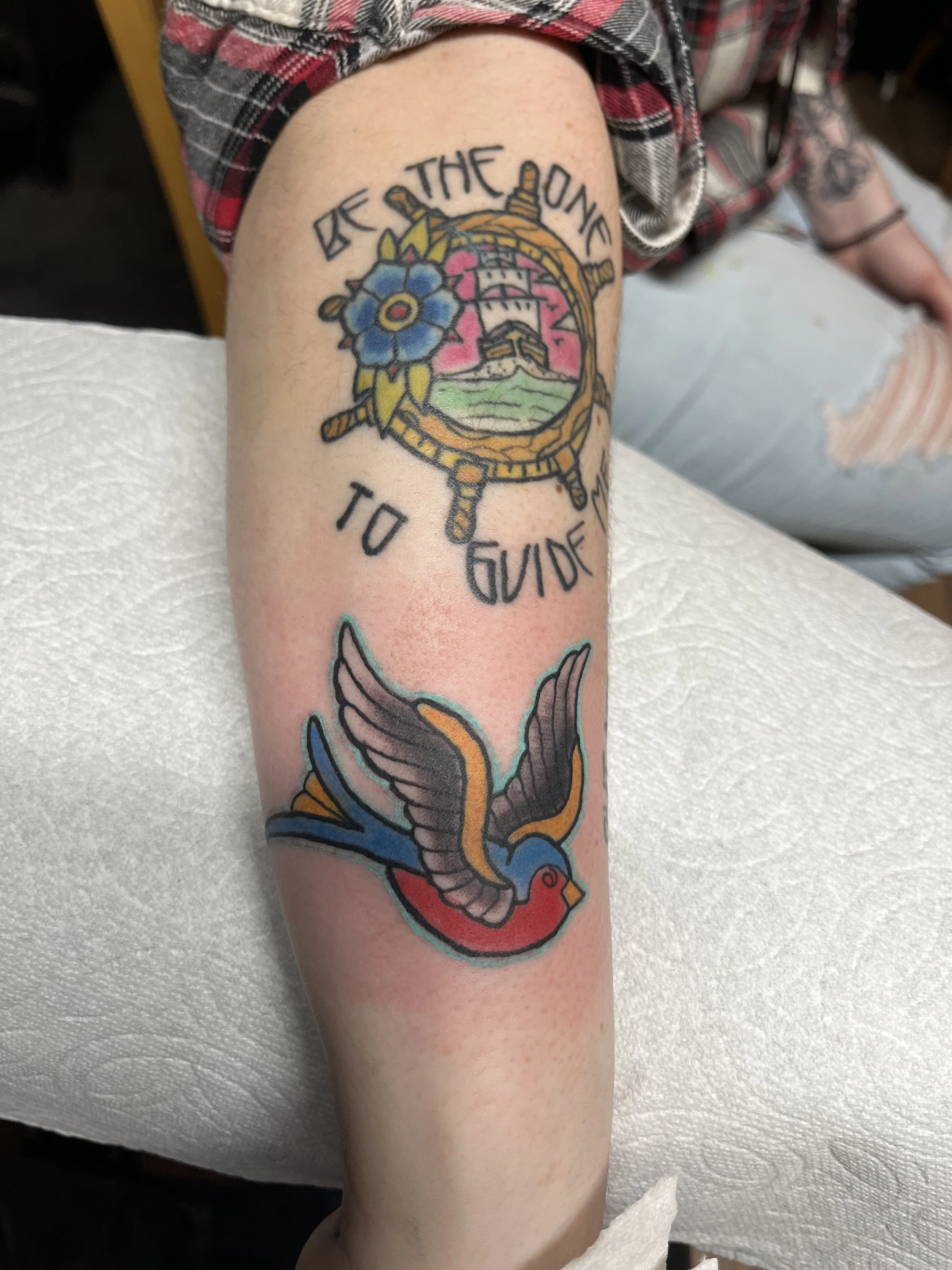 Tattoo on arm featuring a ship's wheel with a blue flower and a lighthouse inside, with the text "BE THE ONE TO GUIDE" above and below. Below the tattoo is a colorful swallow in flight.