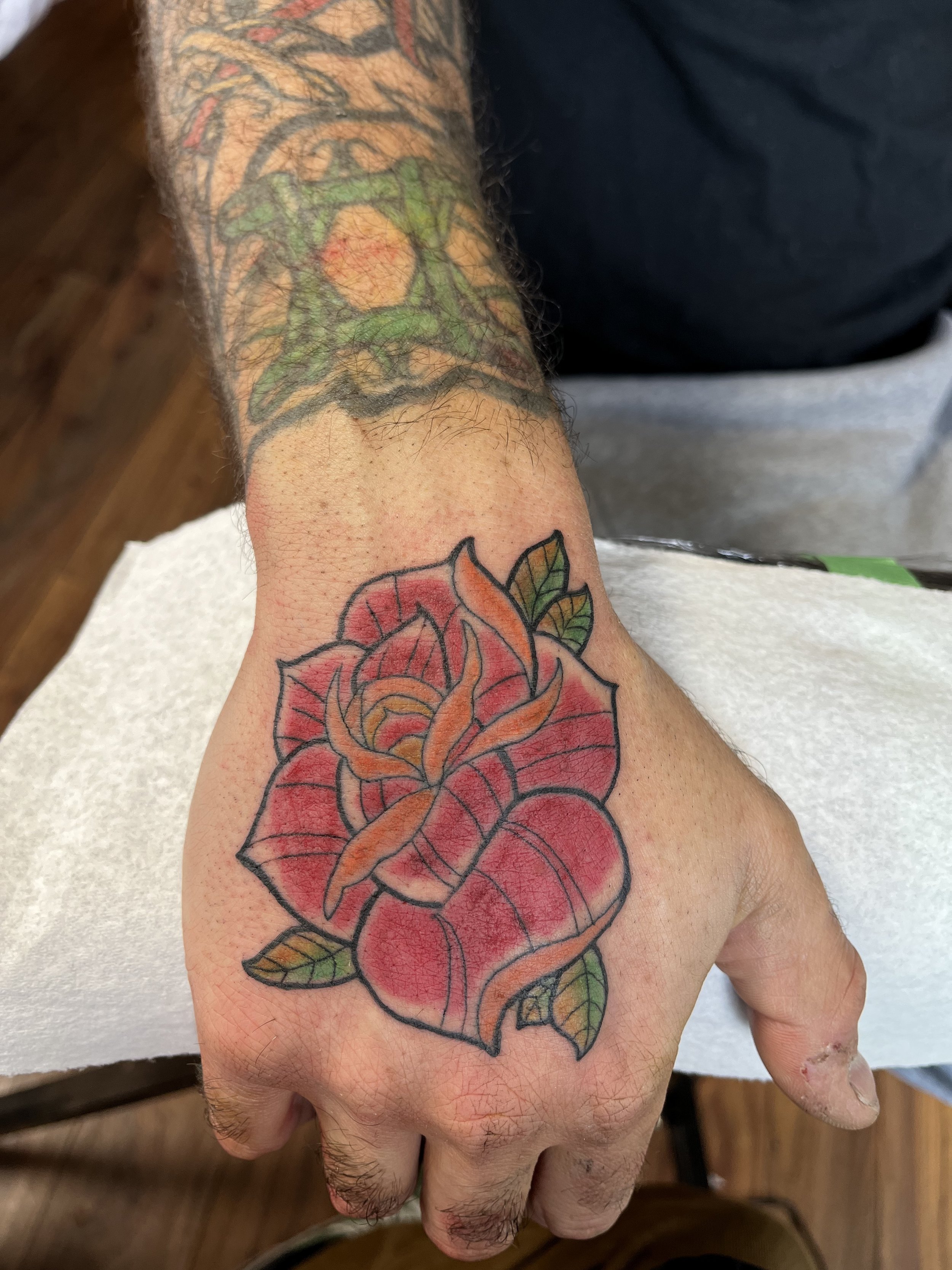 Colorful tattoo of a red rose with orange and yellow accents on the back of a person's hand, with green leaves and black outlines.