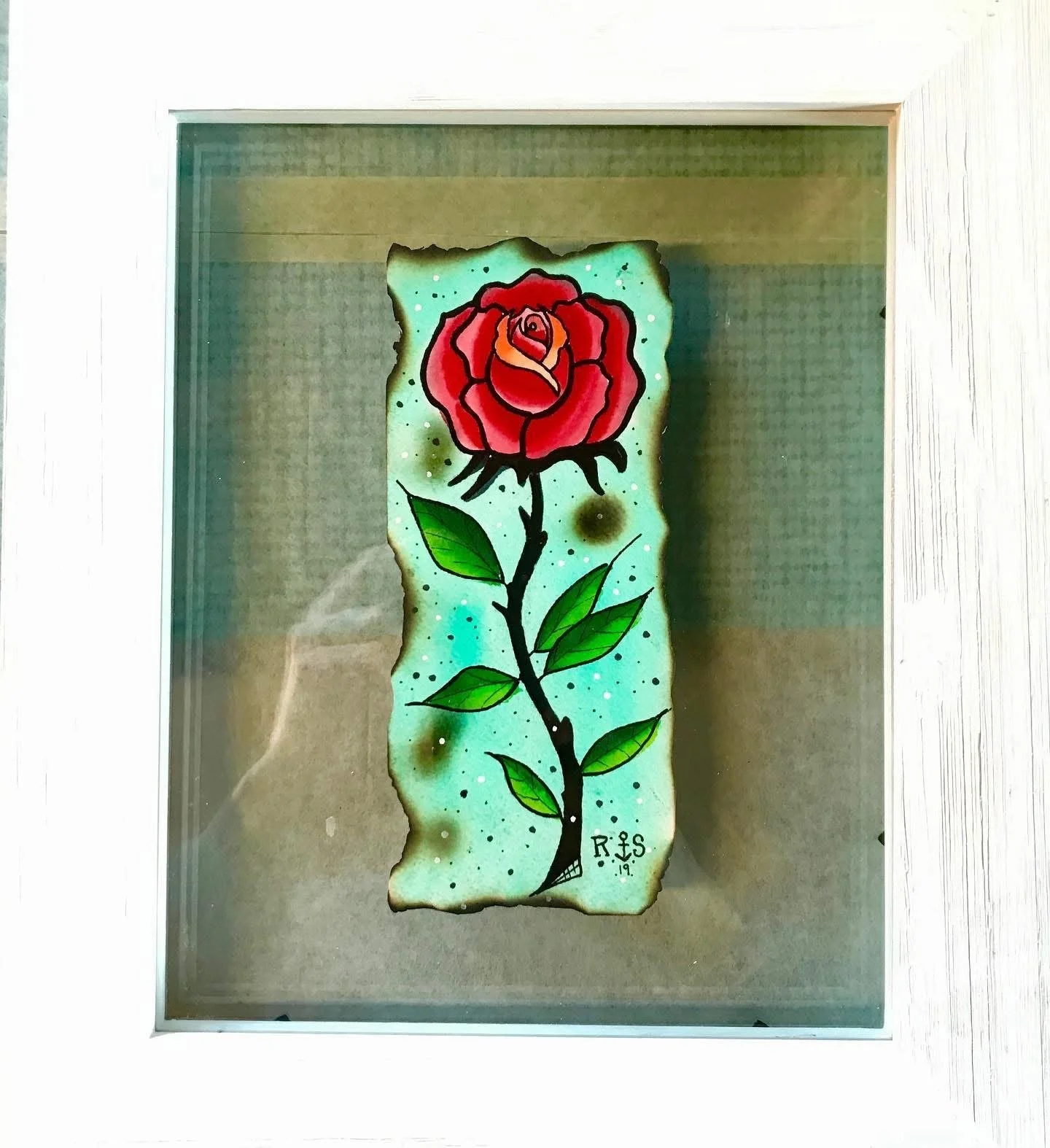 A colorful tattoo-style illustration of a red rose with green leaves on a piece of burnt parchment paper displayed inside a glass case.